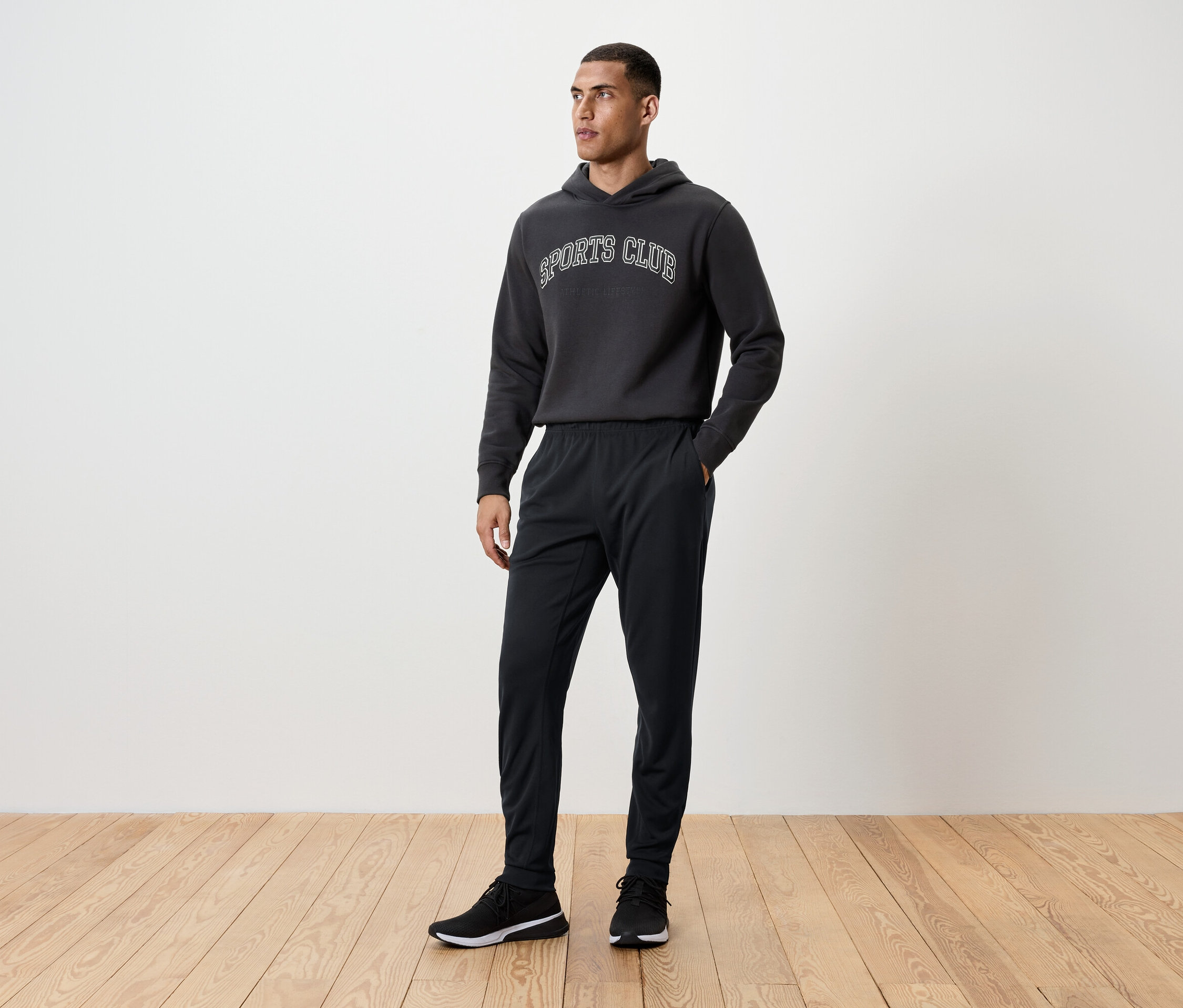 Mężczyzna stoi na drewnianej podłodze, ubrany w czarne spodnie dresowe, bluzę z kapturem i buty sportowe.