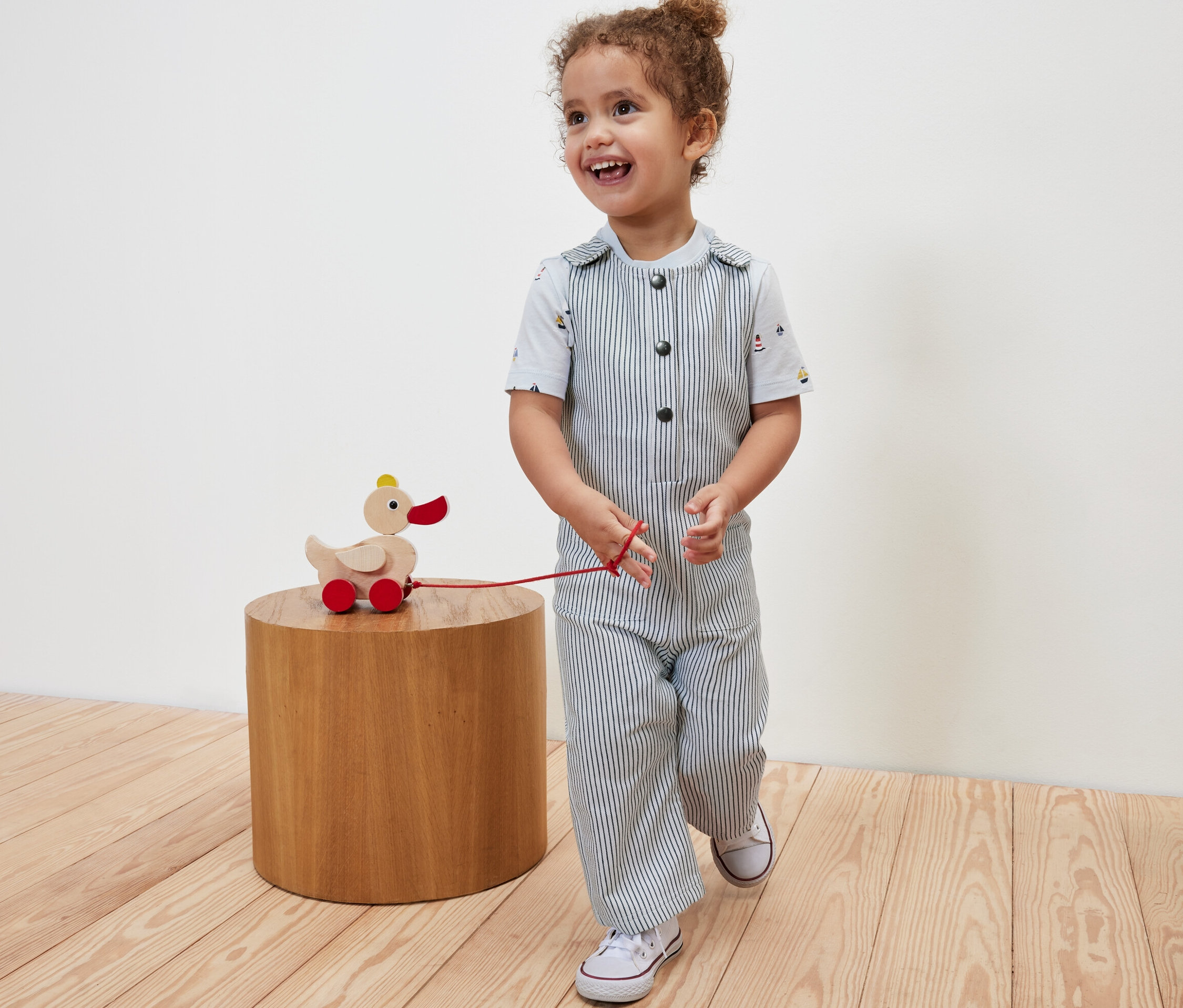 Uśmiechnięte dziecko ciągnie drewnianą zabawkę kaczkę na kółkach i ma na sobie ogrodniczki z tkaniny.