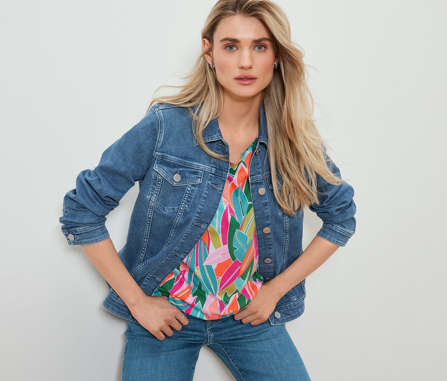 Kobieta pozuje w kurtce dżinsowej, bluzce z obniżoną linią ramion ze wzorem i prostych jeansach.