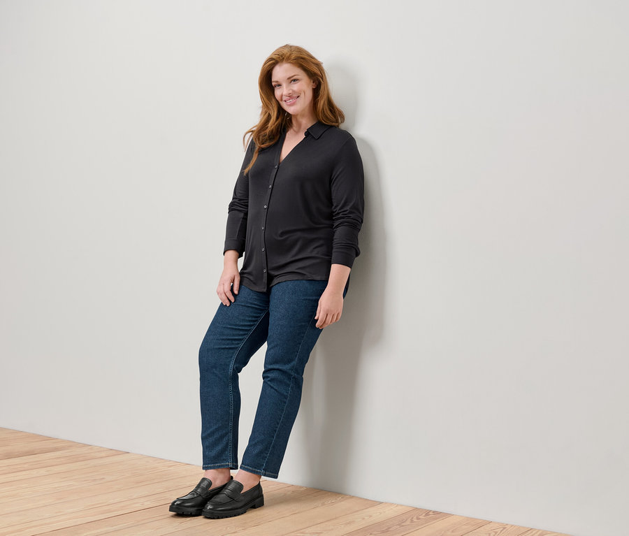 Kobieta z długimi rudymi włosami opiera się o ścianę w czarnym swetrze, dżinsach i czarnych mokasynach.