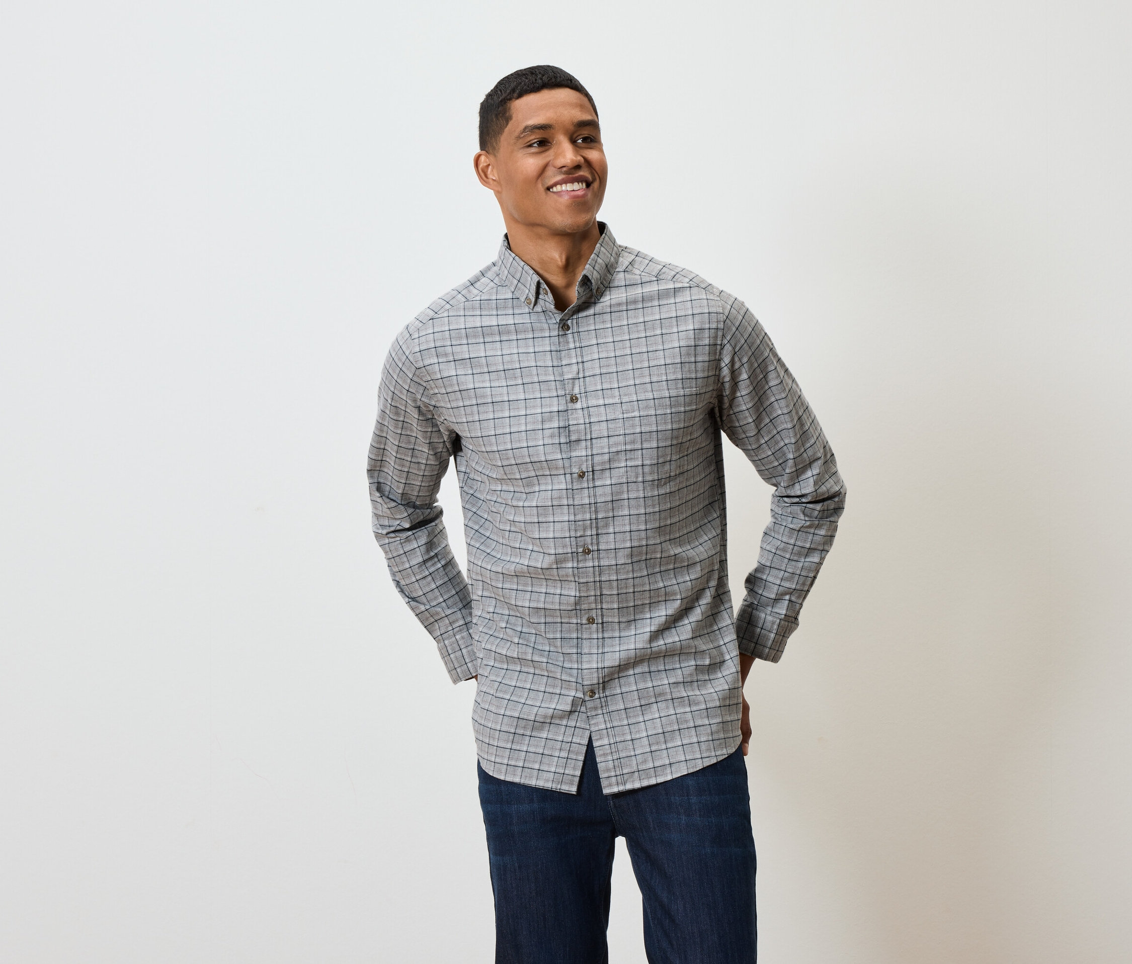 Uśmiechnięty mężczyzna pozuje w szarej koszuli w kratę z długim rękawem i granatowych dżinsach.