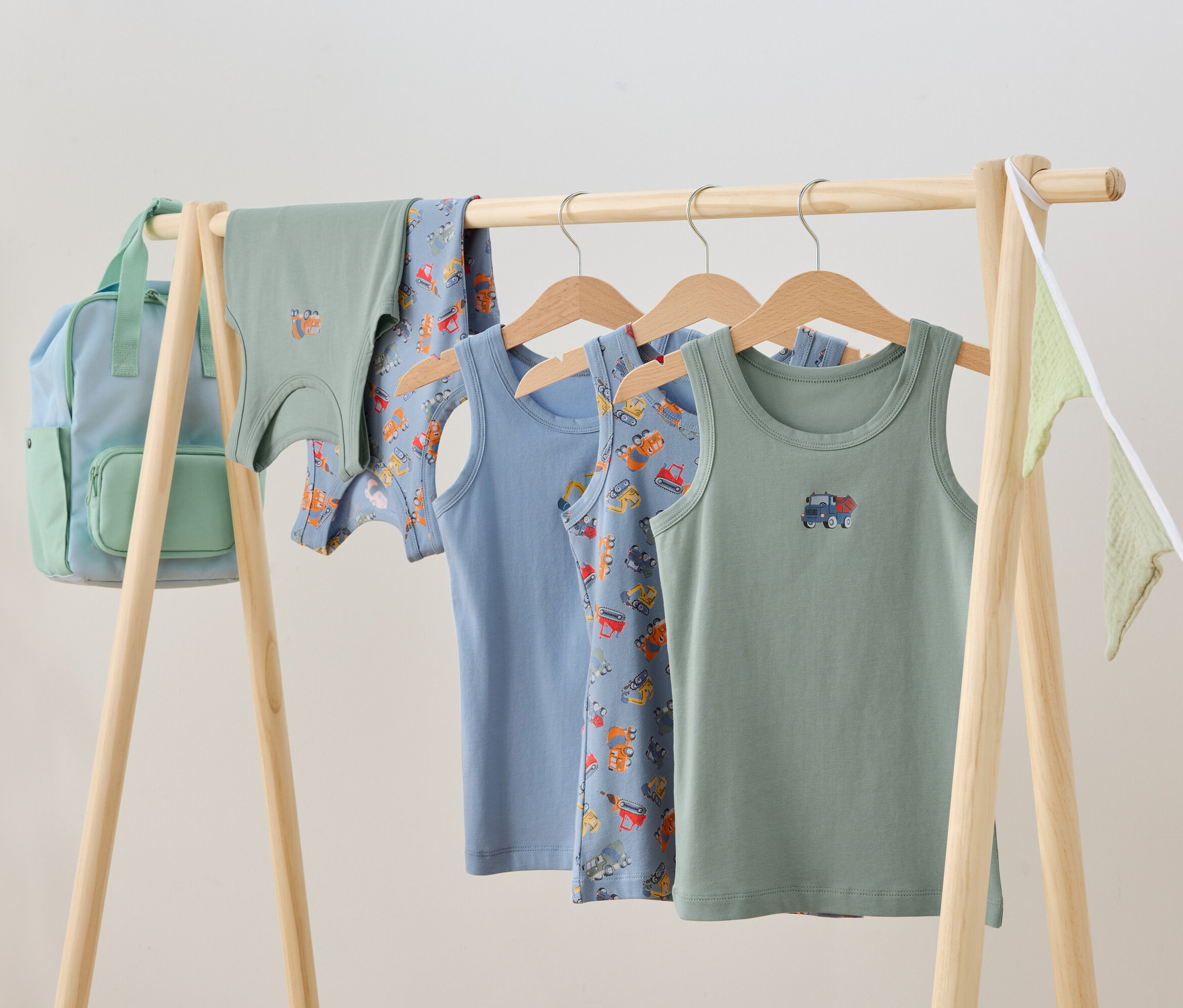 Ubrania dziecięce wiszą na drewnianym stojaku, w tym podkoszulki i plecak.