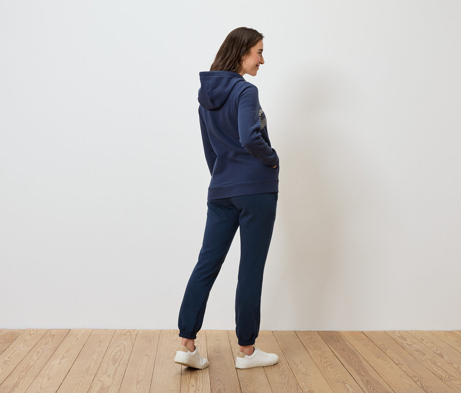 Kobieta w granatowej bluzie z kapturem i spodniach dresowych stoi bokiem na drewnianej podłodze.