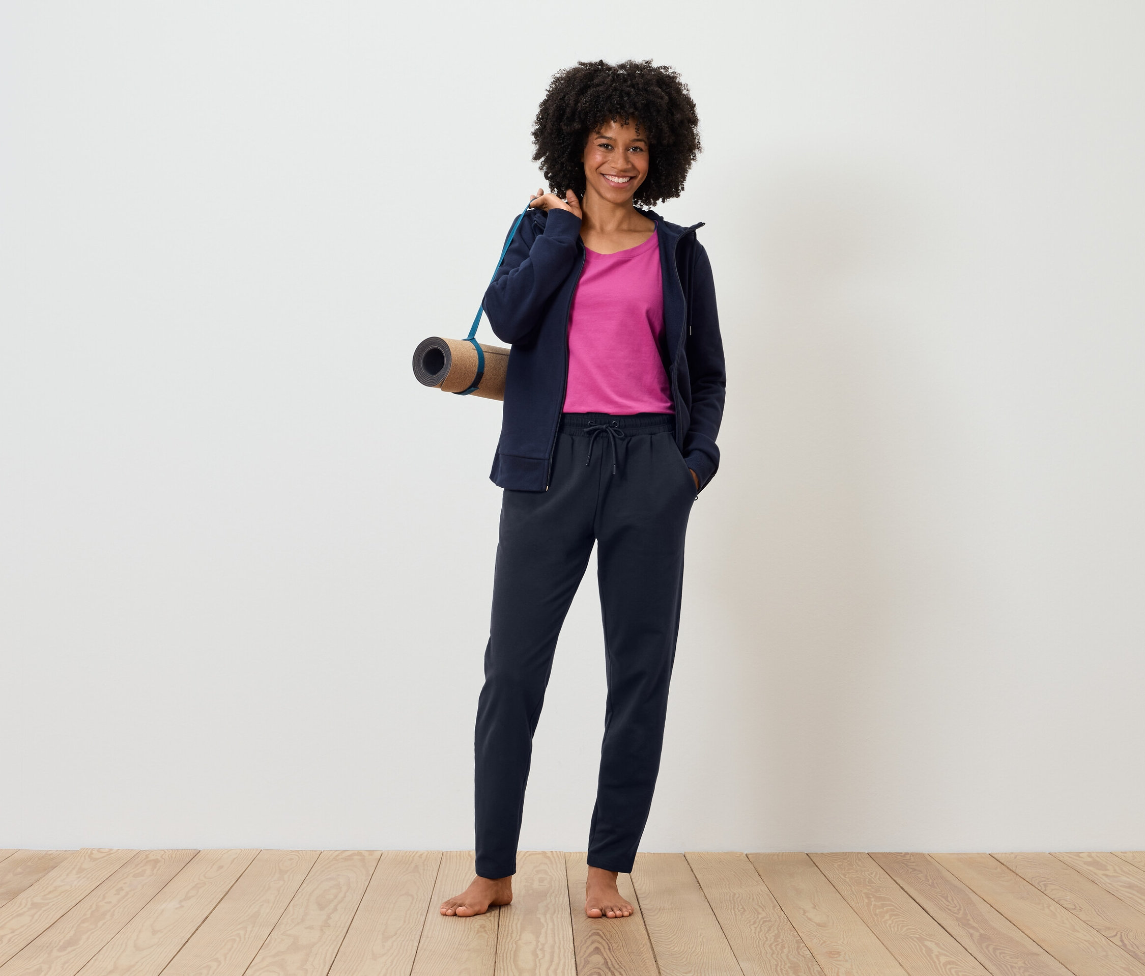 Kobieta stoi boso na drewnianej podłodze, ubrana w bluzę z kapturem i spodnie dresowe, z matą do jogi przewieszoną przez ramię.