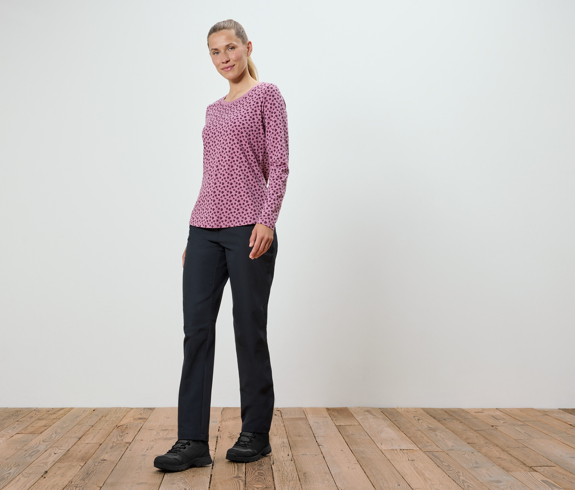 Kobieta w różowej bluzce z długim rękawem i czarnych spodniach stoi na drewnianej podłodze.