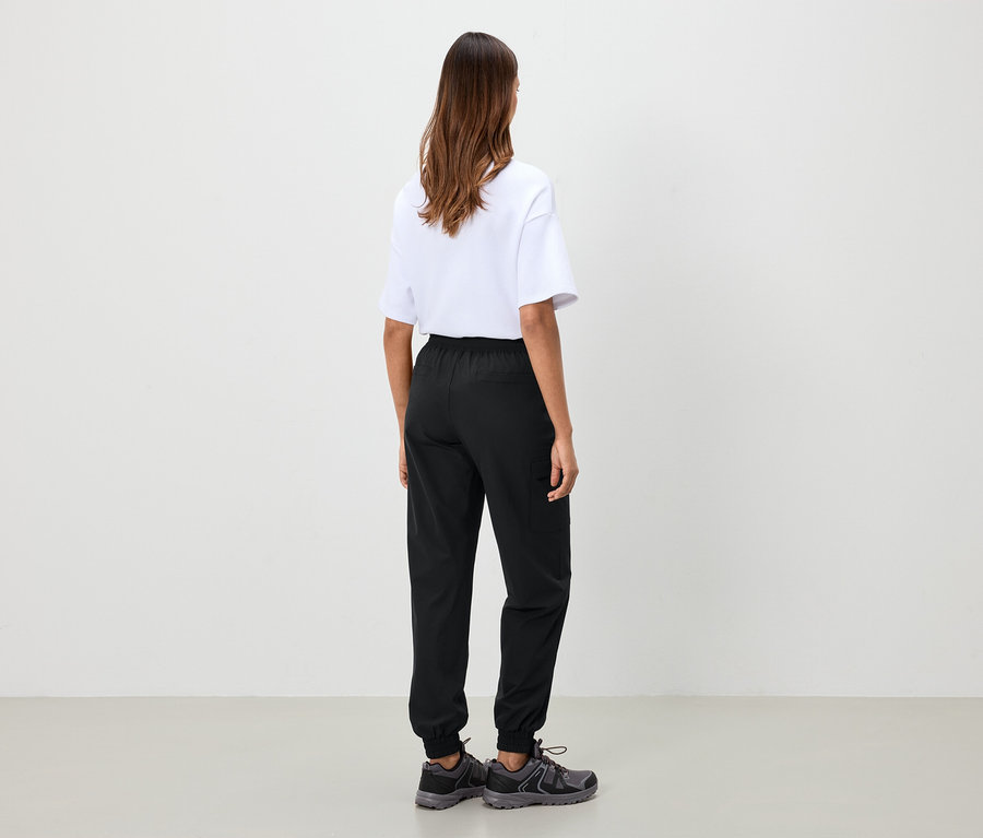 Kobieta stoi tyłem, ubrana w białą koszulkę, czarne spodnie i szare buty.