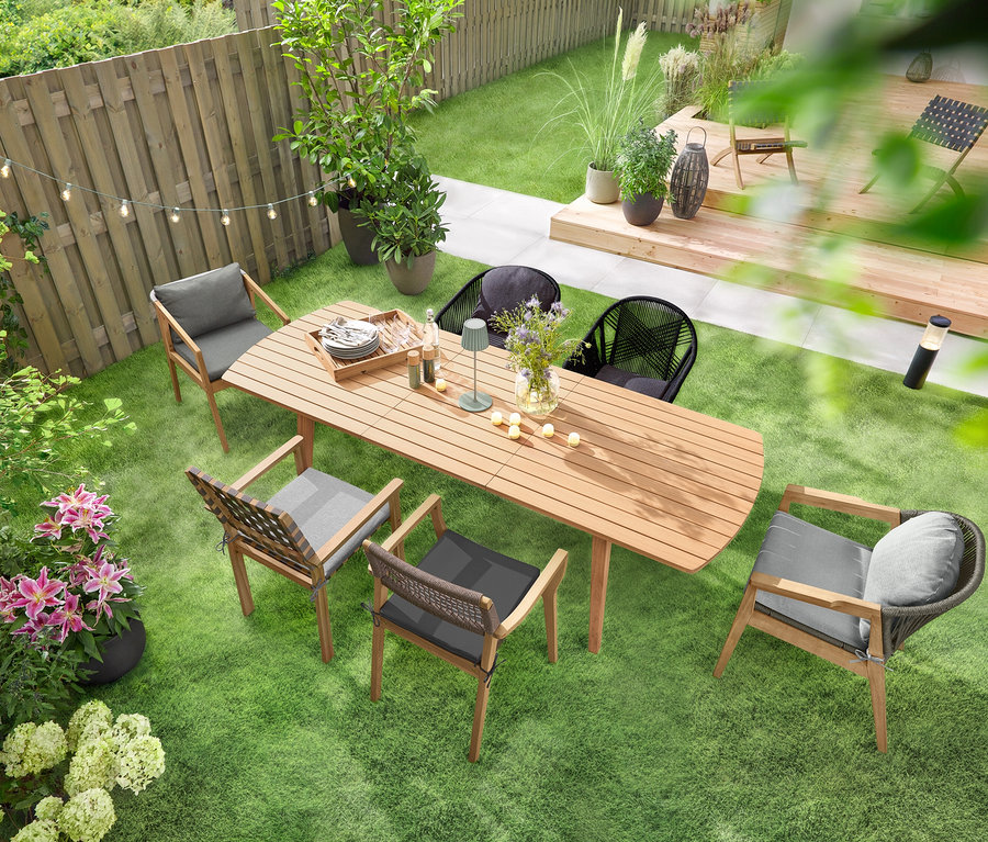 Rozsuwany stół ogrodowy z zaokrąglonymi końcami stoi na zielonym trawniku, wokół niego ustawione są krzesła ogrodowe. Nad stołem wisi łańcuch świetlny LED „Żarówki”, a na nim stoi akumulatorowa lampa stołowa LED do użytku na zewnątrz, szałwiowa. Na stole rozłożonych jest kilka akumulatorowych lampek LED do użytku na zewnątrz, 6 sztuk.