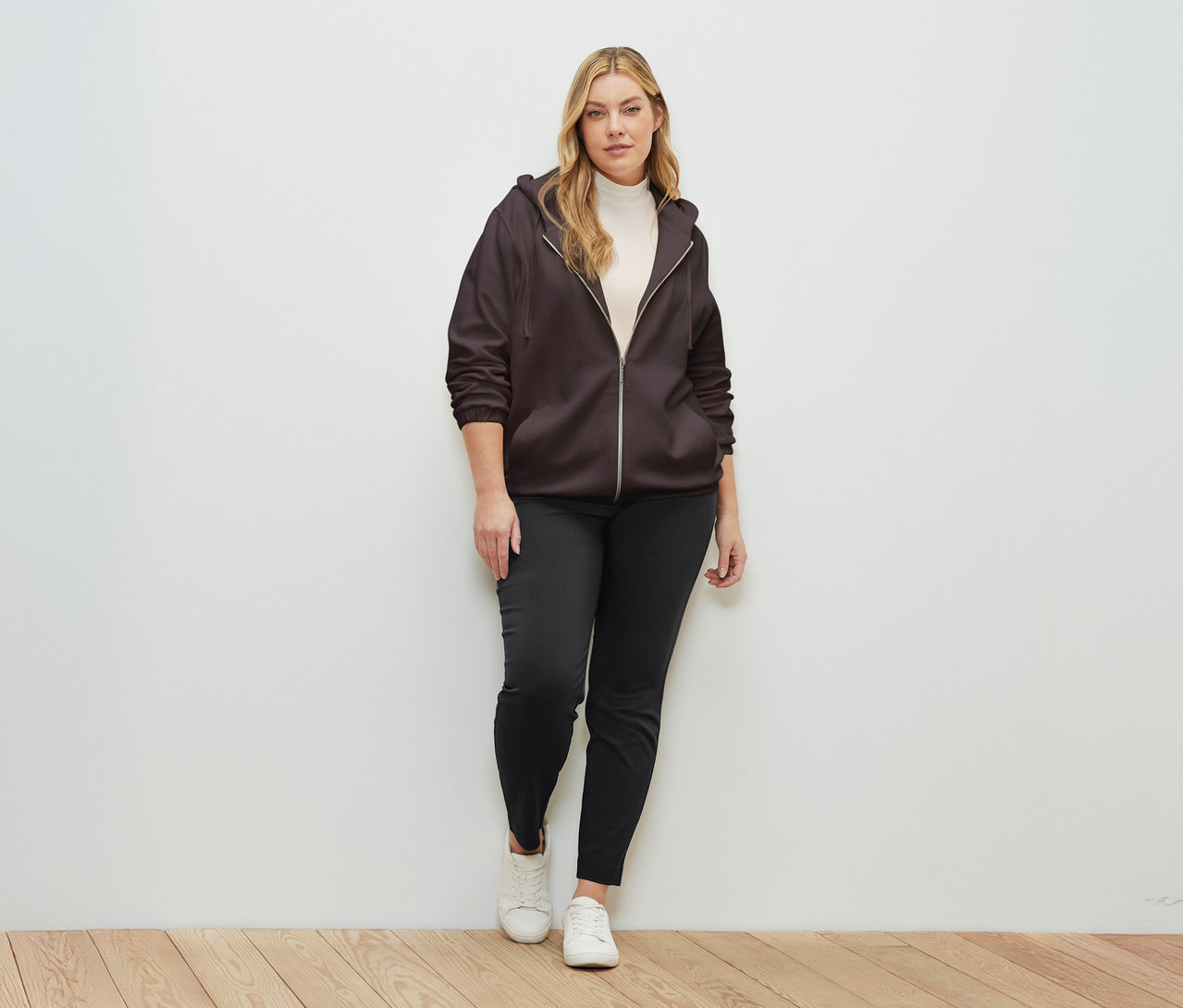 Kobieta stoi na tle ściany, ubrana w brązową rozpinaną bluzę dresową.