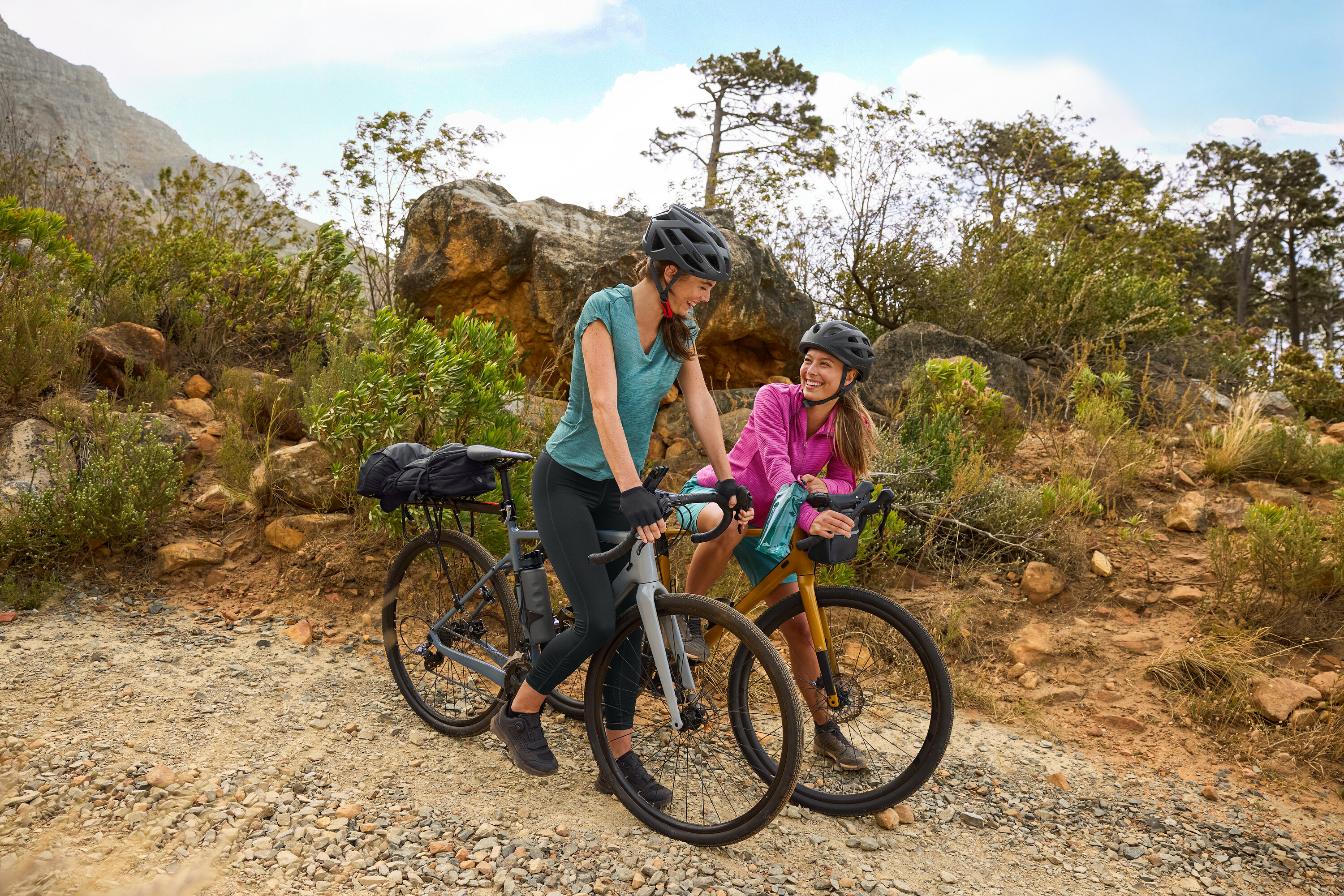 Dwie kobiety w kaskach stoją z rowerami na żwirowej drodze w pagórkowatym terenie. Bidon przymocowany jest do roweru.