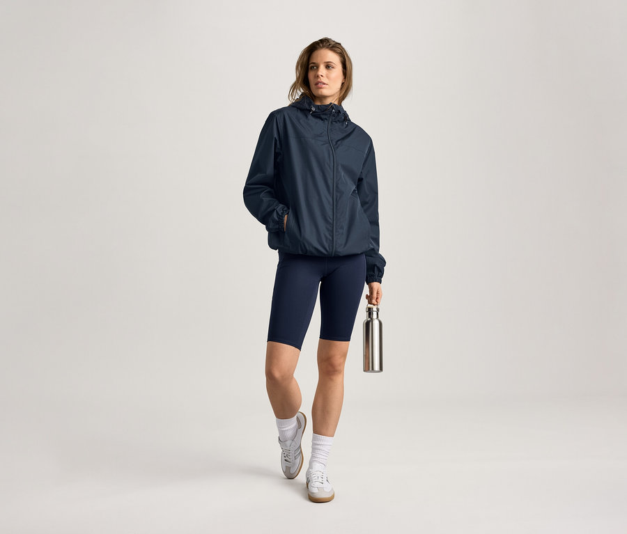 Kobieta w kurtce przeciwdeszczowej, kolarkach i butelce na wodę. Ma na sobie skarpetki i tenisówki.