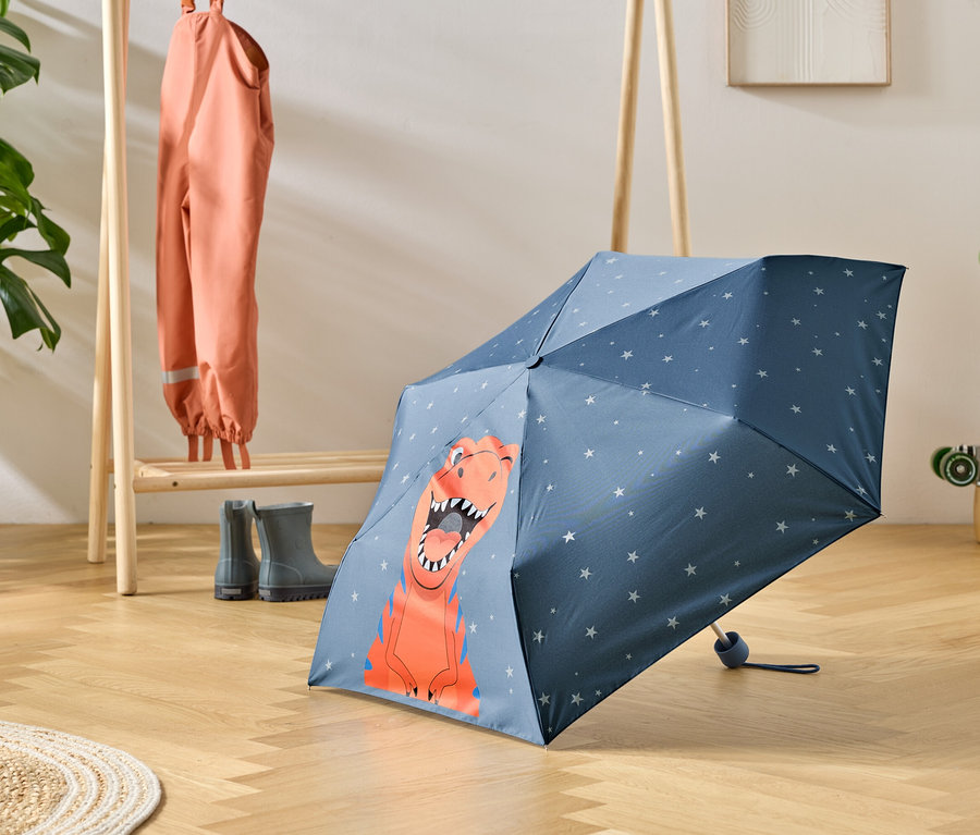 Niebieski parasol w dinozaury stoi na drewnianej podłodze, obok wieszak z ubraniami przeciwdeszczowymi i kaloszami.