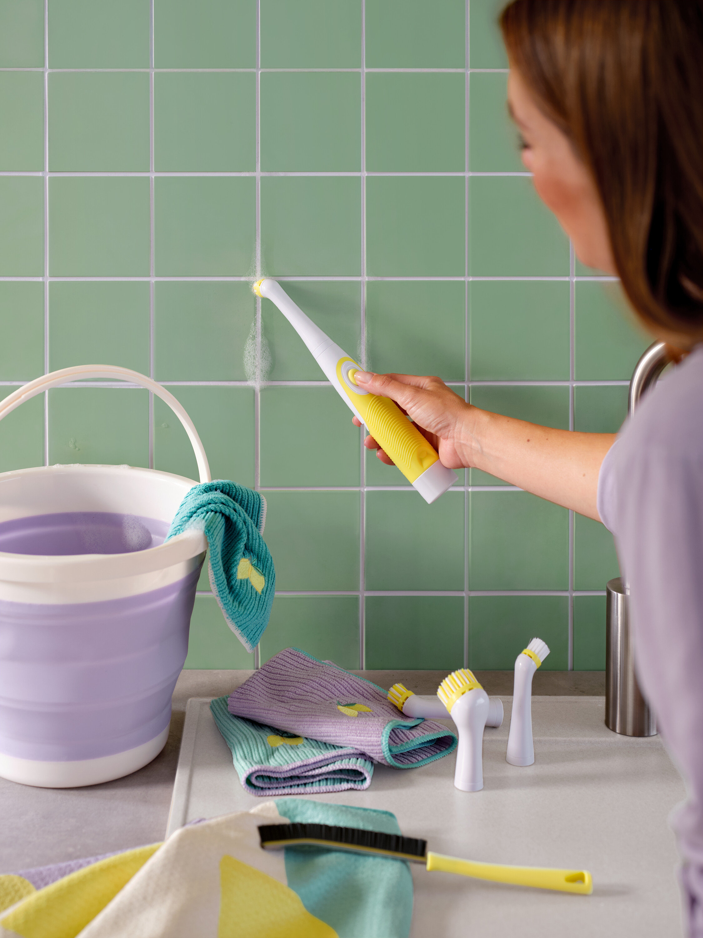 Kobieta czyści zielone kafelki żółto-białą szczotką, widoczne jest również wiadro i ściereczki.