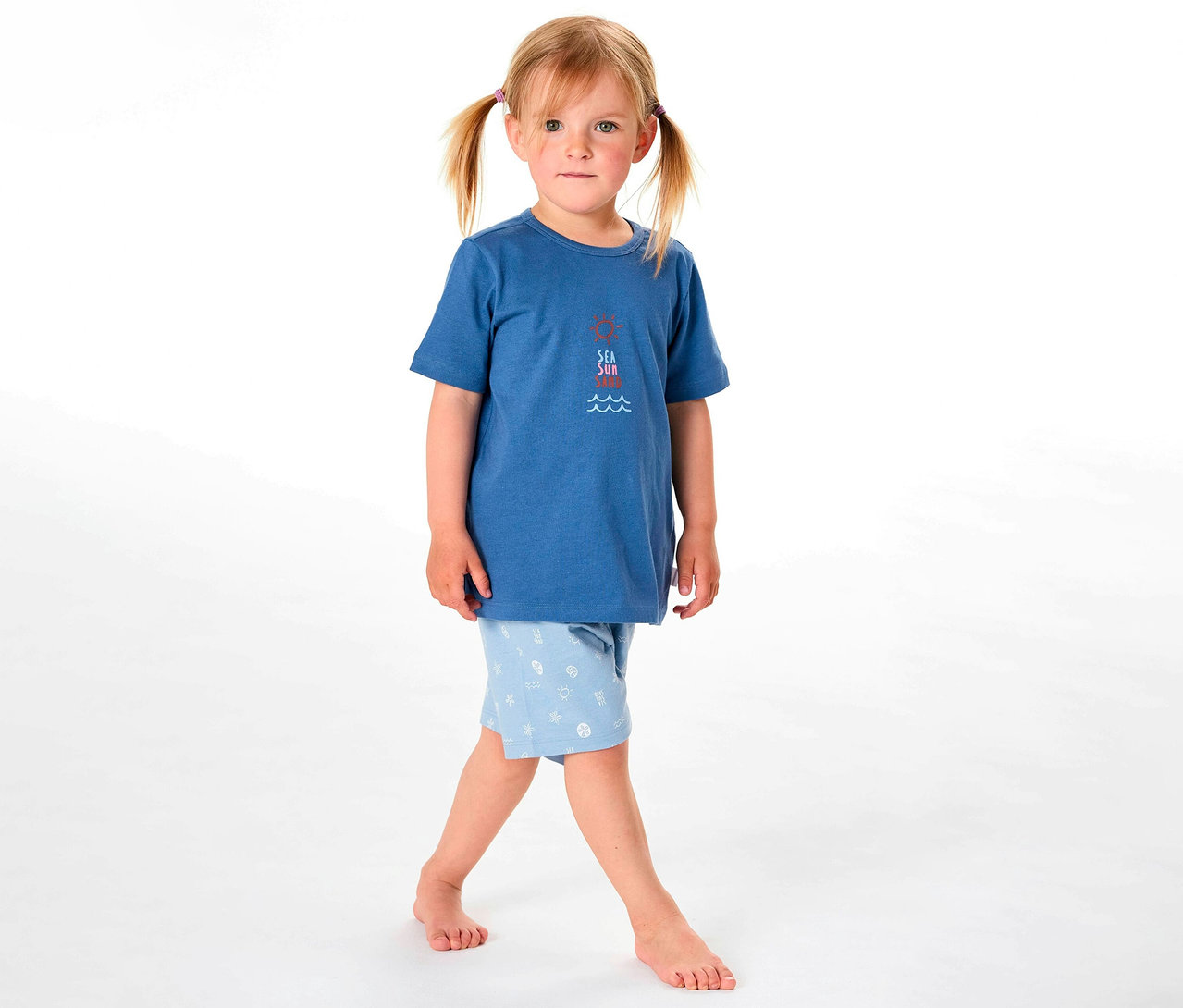 Dziewczynka ma na sobie piżamę dziecięcą SCHIESSER: niebieską koszulkę i jasnoniebieskie szorty we wzory.