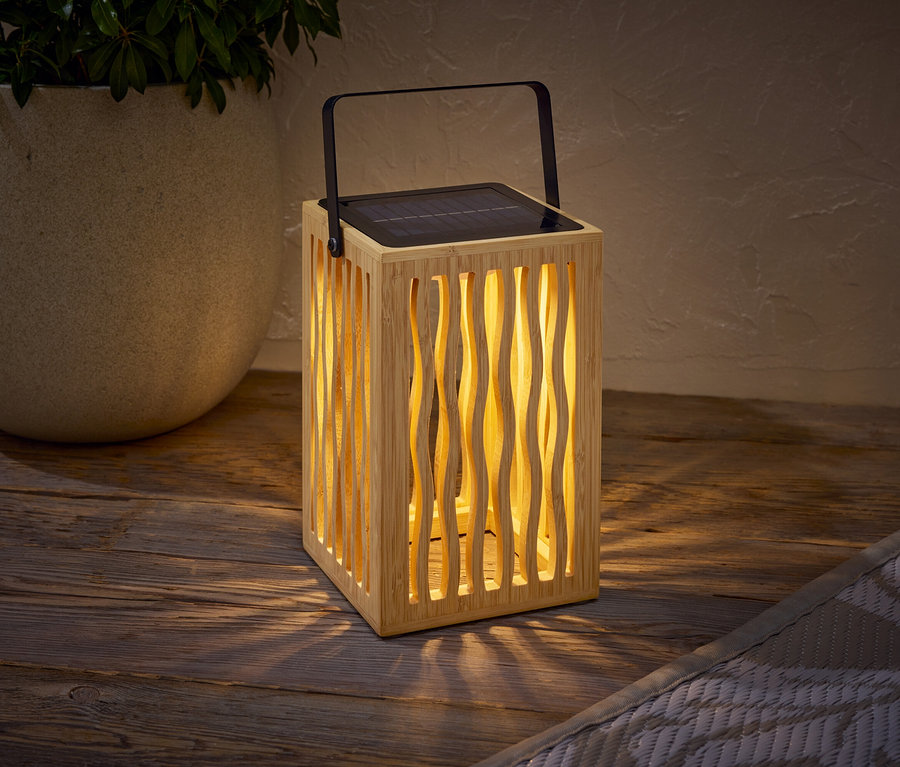 Drewniana lampa solarna z czarną rączką stoi na drewnianym stole obok doniczki z rośliną.