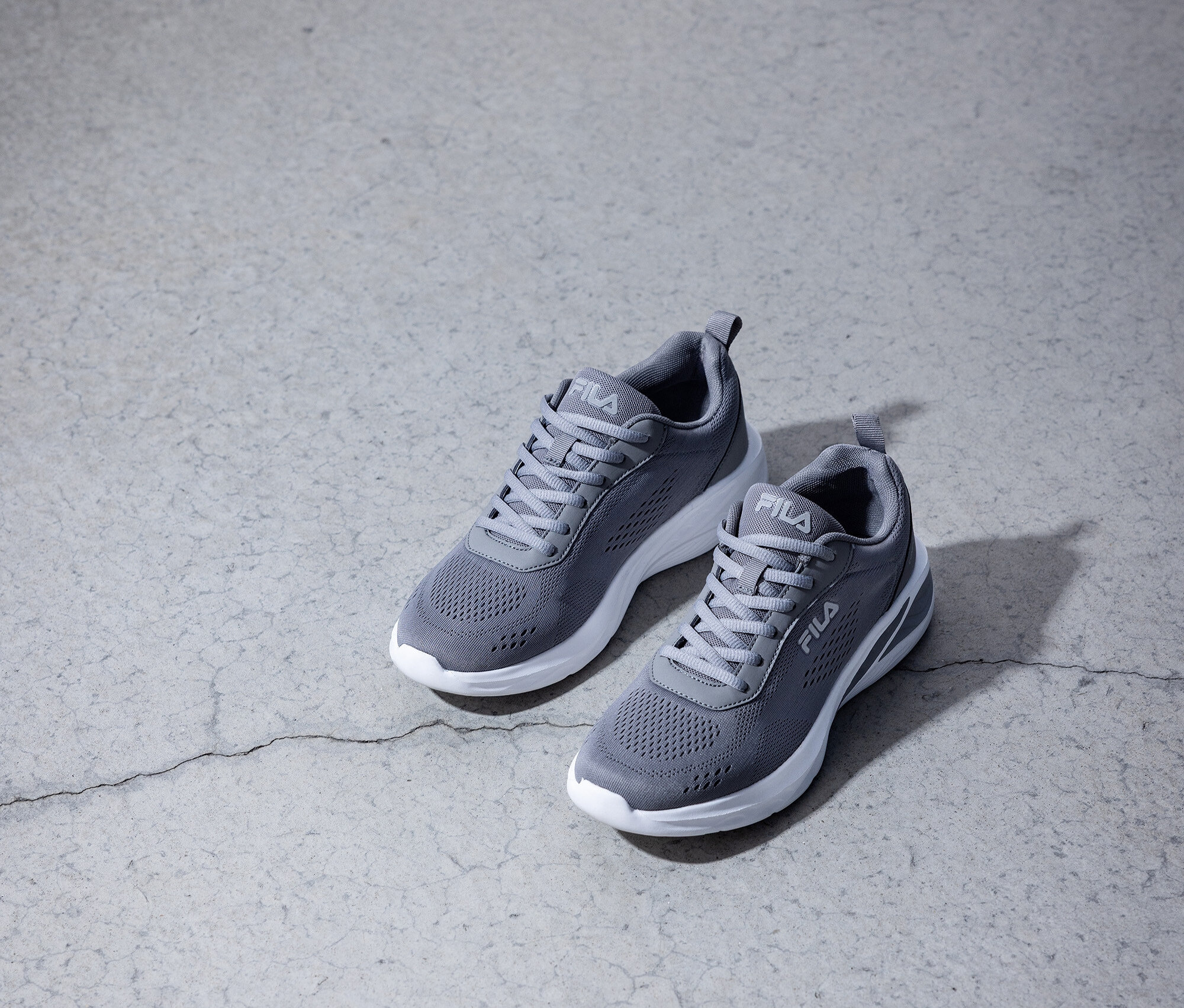 Dwa szare sneakersy Fila z białymi podeszwami stoją na szarej betonowej podłodze.