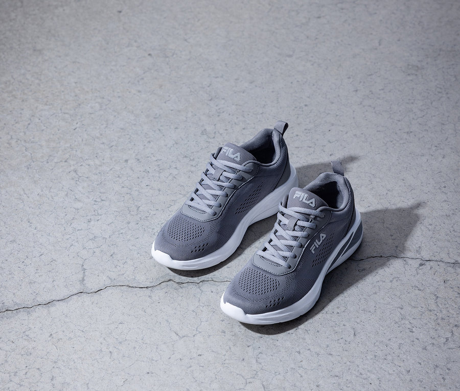 Dwa szare sneakersy Fila z białymi podeszwami stoją na szarej betonowej podłodze.