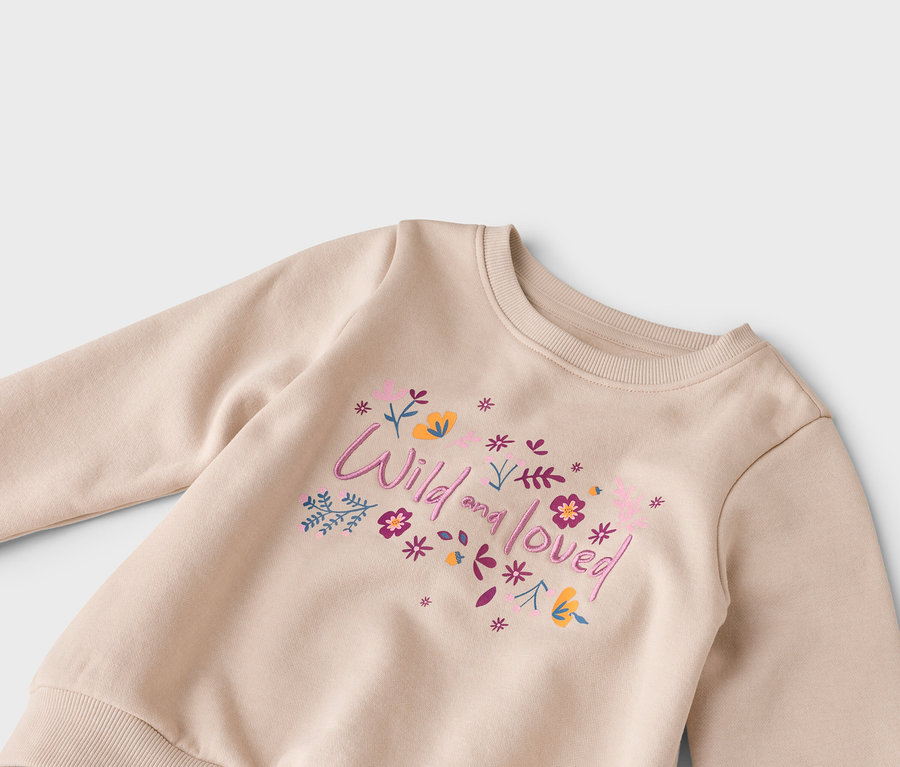 Zbliżenie beżowej bluzy z napisem „Wild and loved” i kwiatami w kolorze różowym, fioletowym i żółtym.