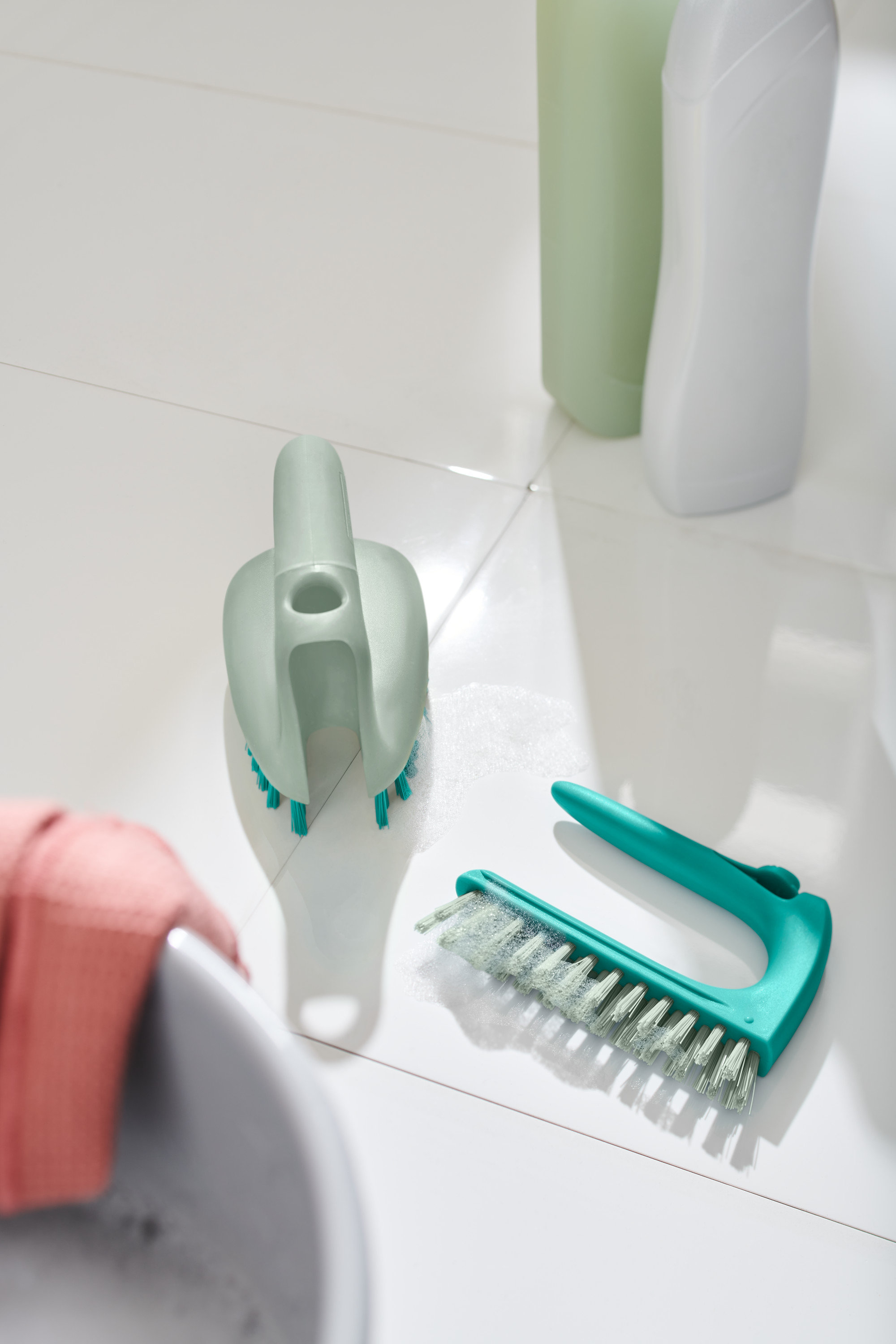 Dwie szczotki do posadzek z płytek 2 w 1 leżą na białych płytkach obok umywalki.