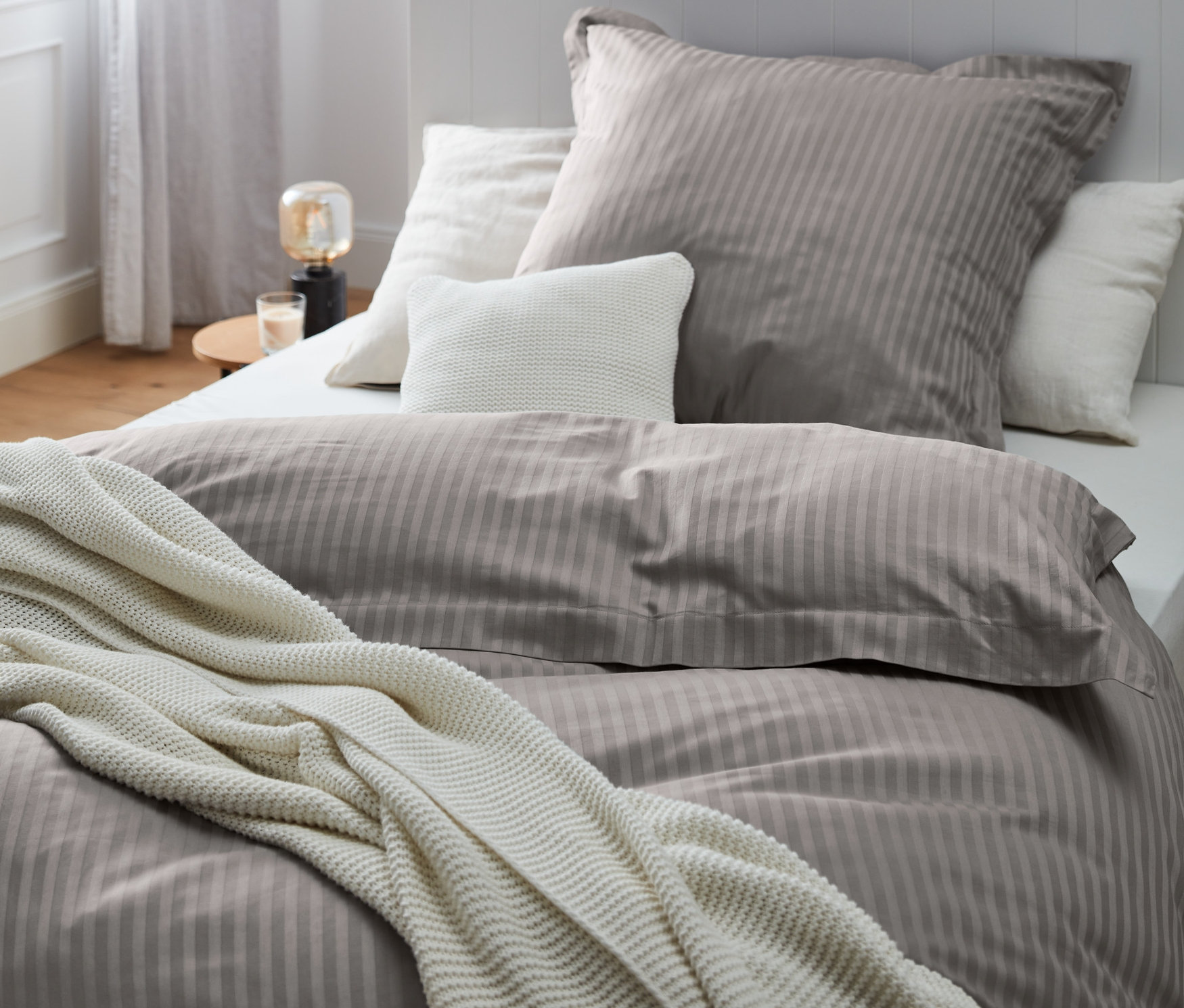 Zbliżenie na łóżko z szarą satynową pościelą premium w różnych rozmiarach i białym kocem z dzianiny.