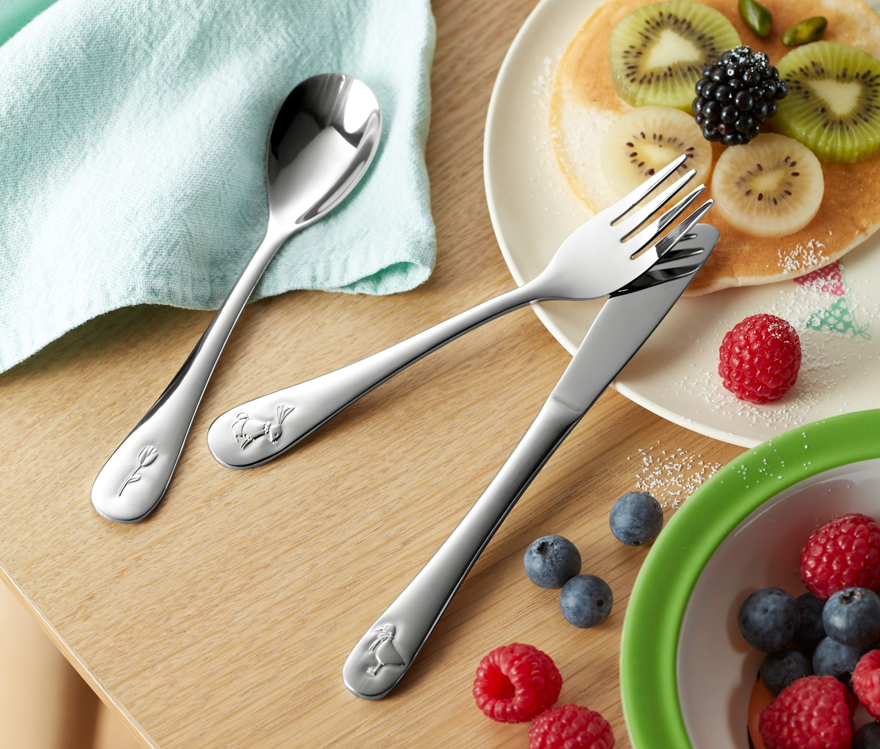 Na drewnianym stole leżą: 3-częściowy zestaw sztućców dla dzieci, talerz z naleśnikami i owocami, miska z jagodami i malinami oraz szklanka z napojem.