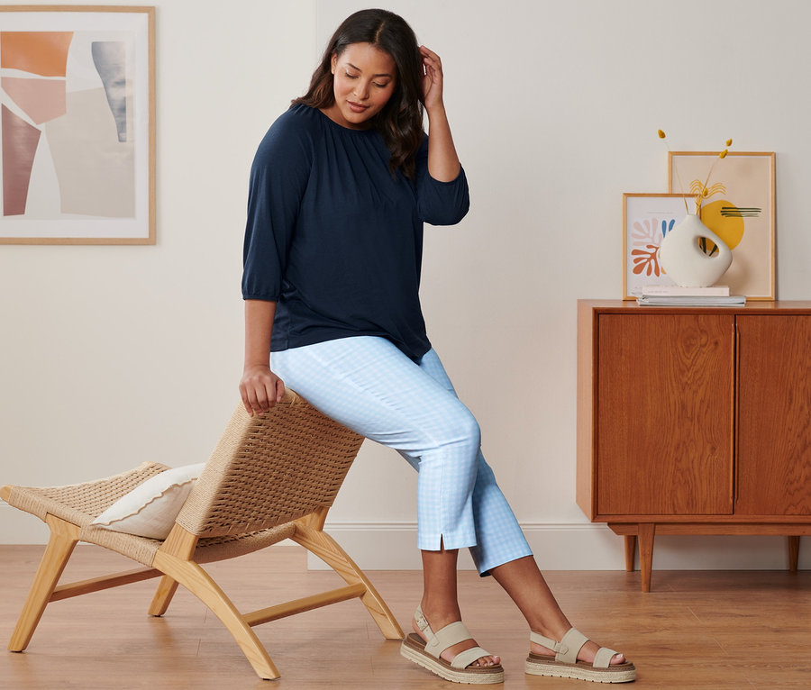 Kobieta siedzi na krześle i ma na sobie granatową bluzkę z rękawami o długości 3/4, błękitno-białe spodnie stretchowe o długości 7/8 i skórzane sandały na platformie.