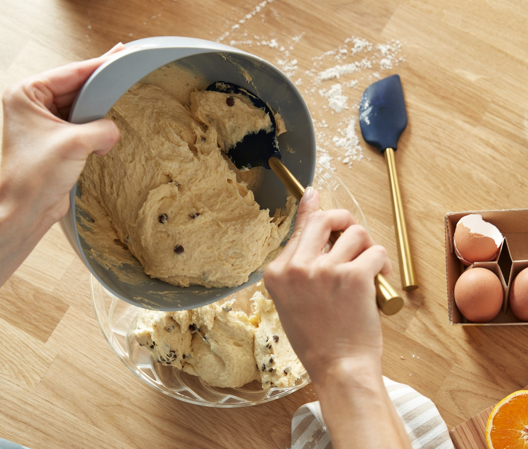Kobieta przelewa ciasto z czekoladą z szarej miski do szklanej miski. Na drewnianym stole leżą 2 skrobaki do ciasta, jajka i pomarańcza.