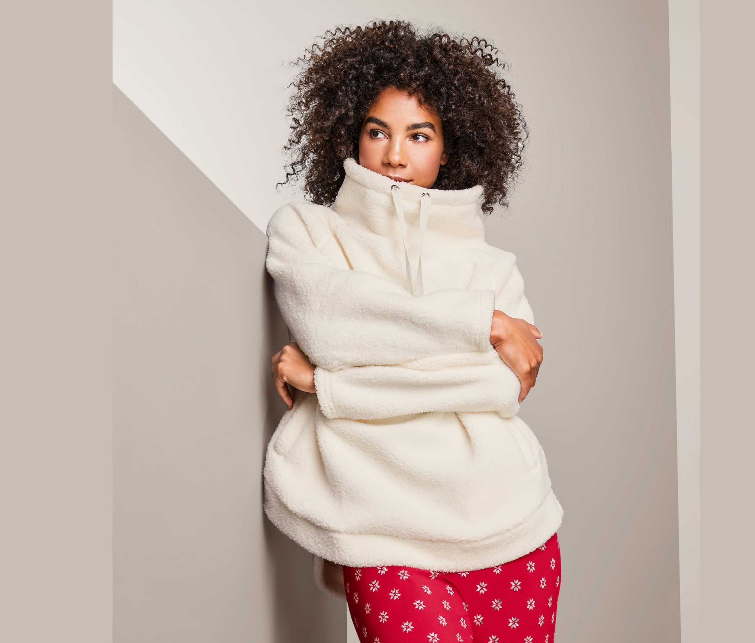 Kobieta o kręconych włosach opiera się o ścianę, ubrana w pluszową bluzę damską i czerwone spodnie.