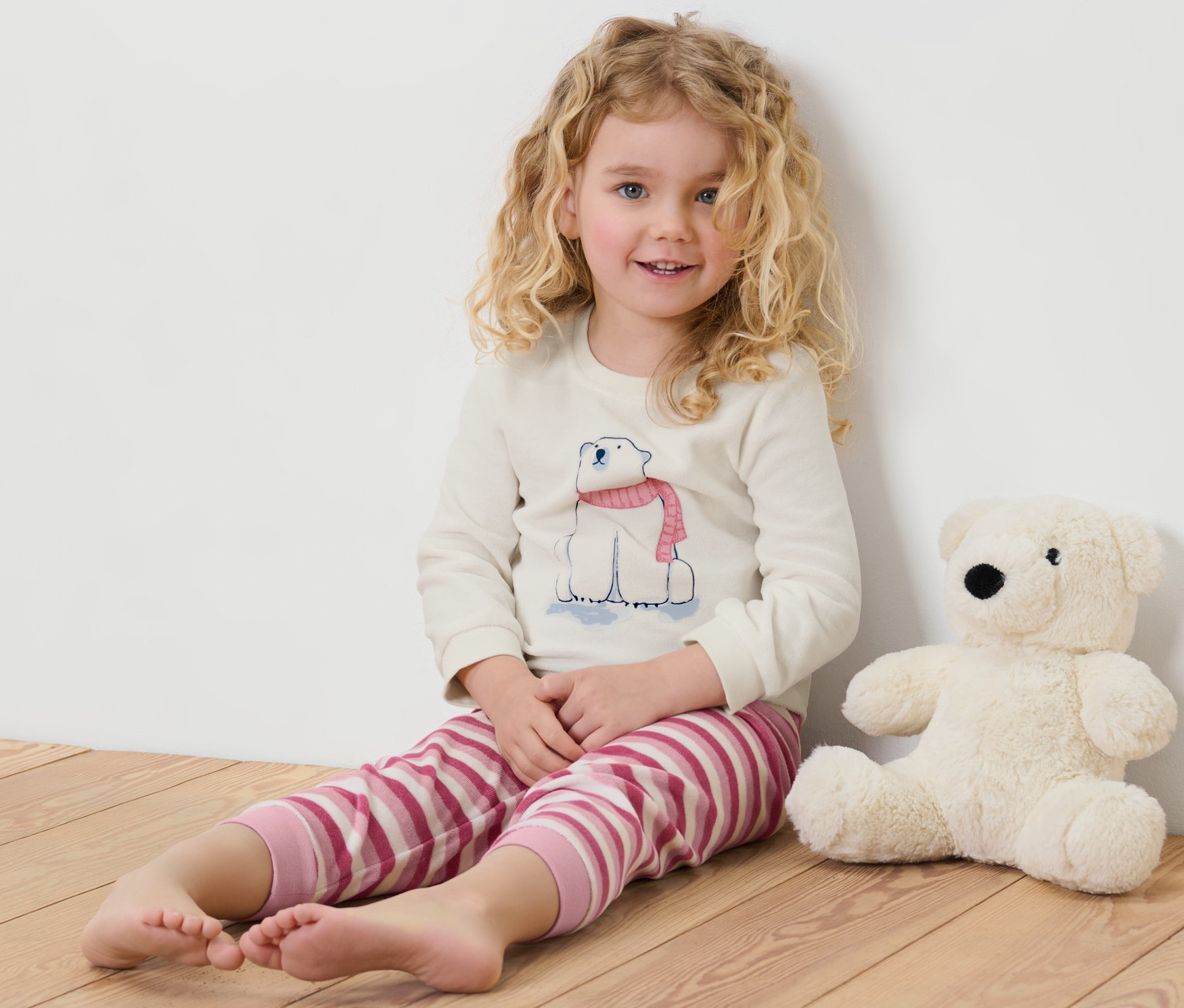 Dziewczynka siedzi na podłodze obok pluszowego misia polarnego. Ma na sobie piżamę dziecięcą z weluru z motywem niedźwiedzia polarnego.