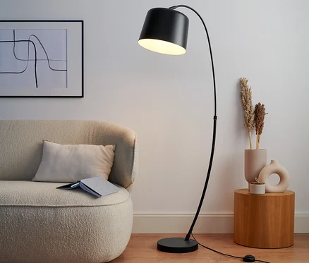 Czarna metalowa lampa stojąca stoi obok beżowej sofy.