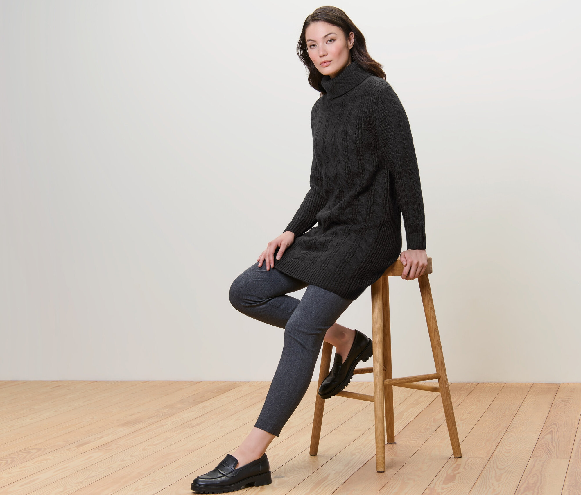 Kobieta siedzi na stołku i ma na sobie długi czarny sweter z warkoczowym wzorem.