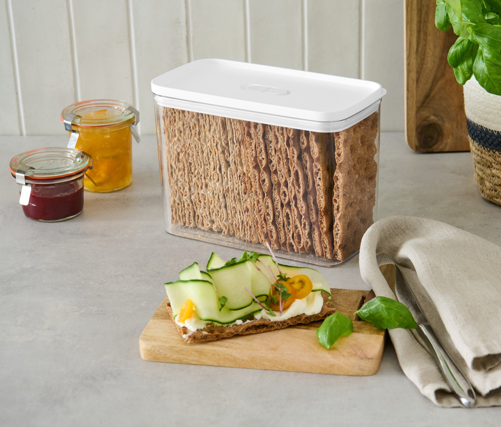 Pojemnik na żywność z białą pokrywką wypełniony krakersami chlebowymi stoi na blacie obok słoików z dżemem.