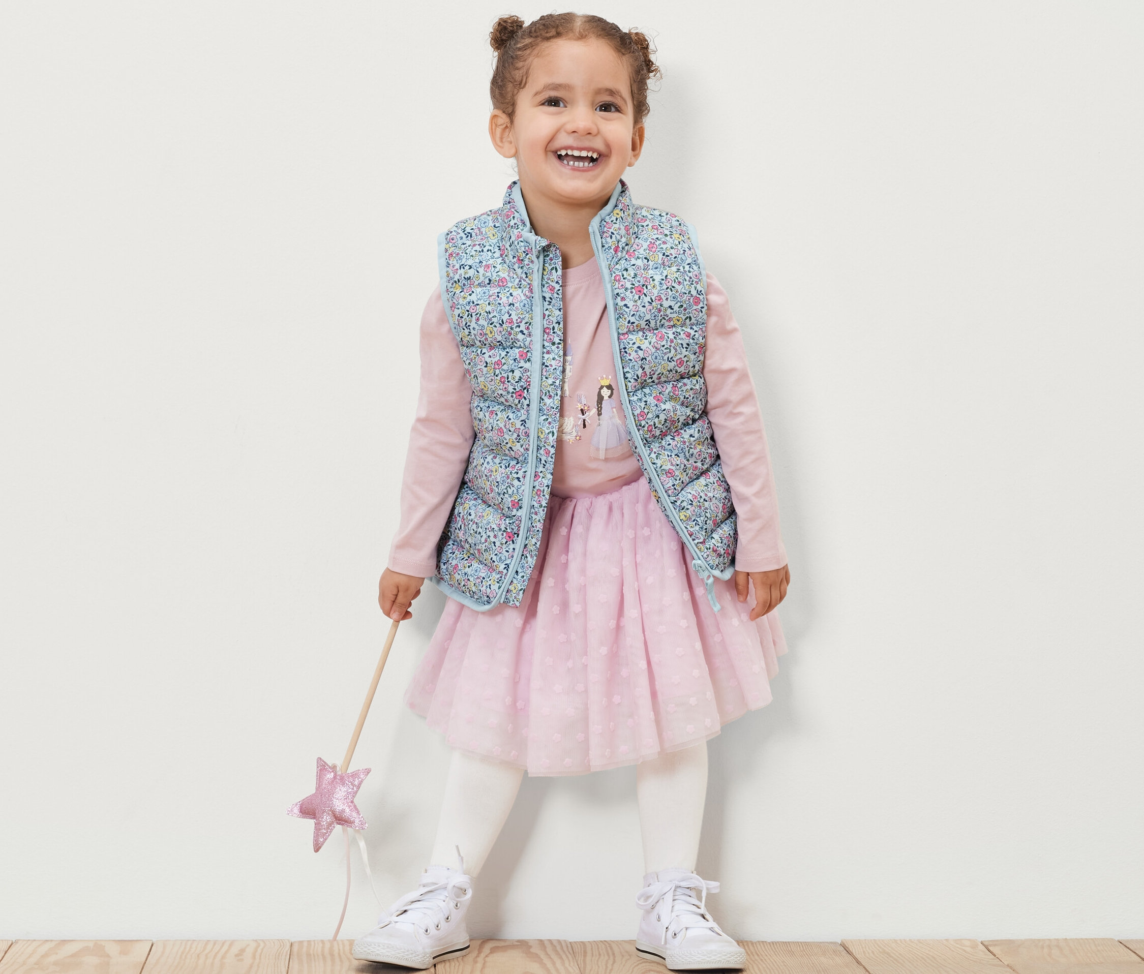 Uśmiechnięta dziewczynka ma na sobie pikowaną kamizelkę w kwiaty, różową bluzkę z długim rękawem, różową spódnicę i białe tenisówki, trzyma w ręku różdżkę.