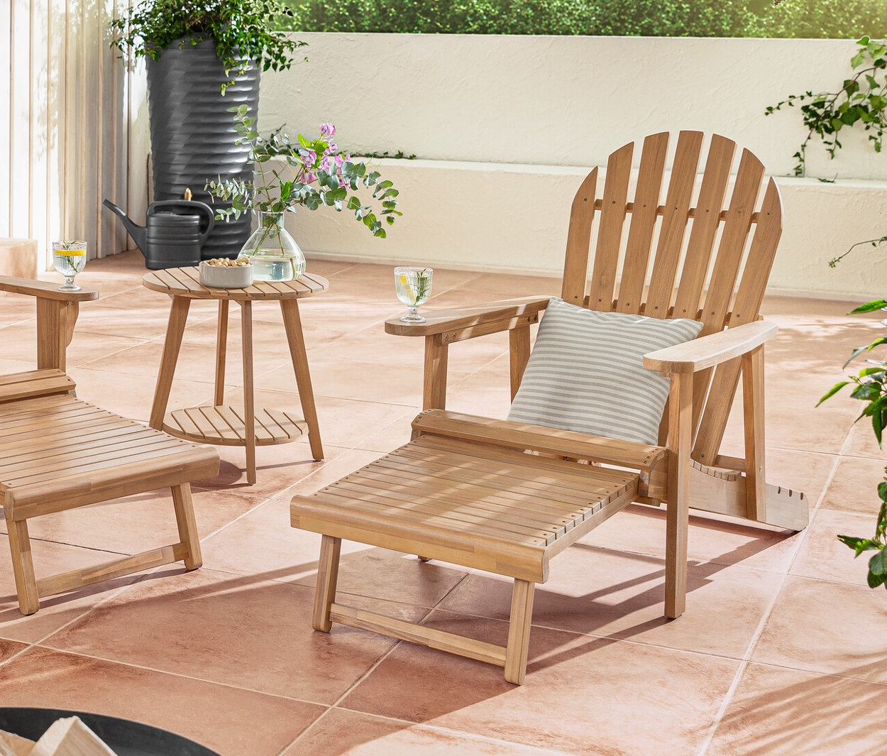 Podwójny i pojedynczy fotel wypoczynkowy typu Adirondack z wysuwanym podnóżkiem stoi na tarasie z poduszkami ogrodowymi o strukturze lnu.