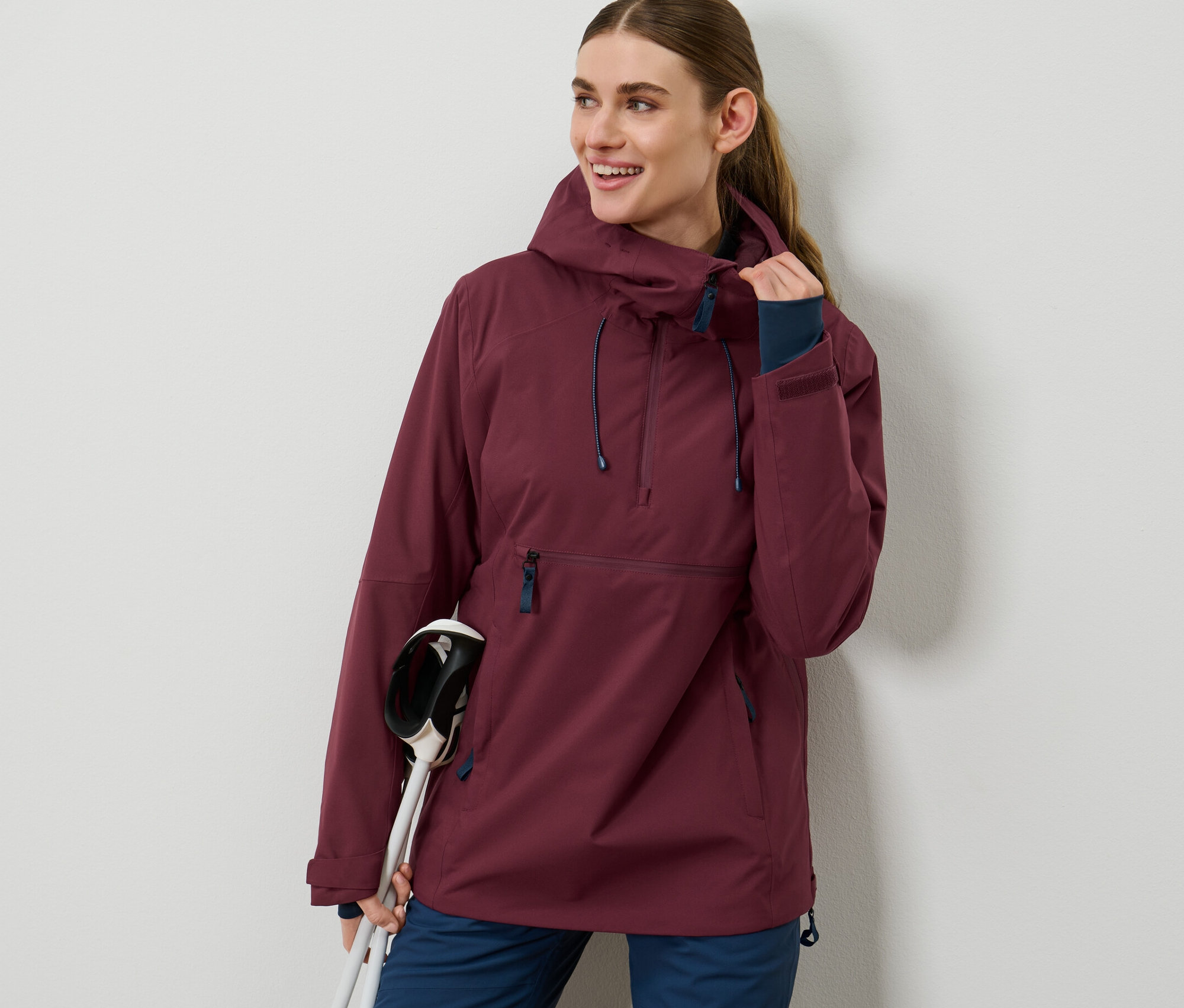Kobieta ma na sobie bordową kurtkę narciarską z krótkim zamkiem błyskawicznym i trzyma kijki narciarskie.