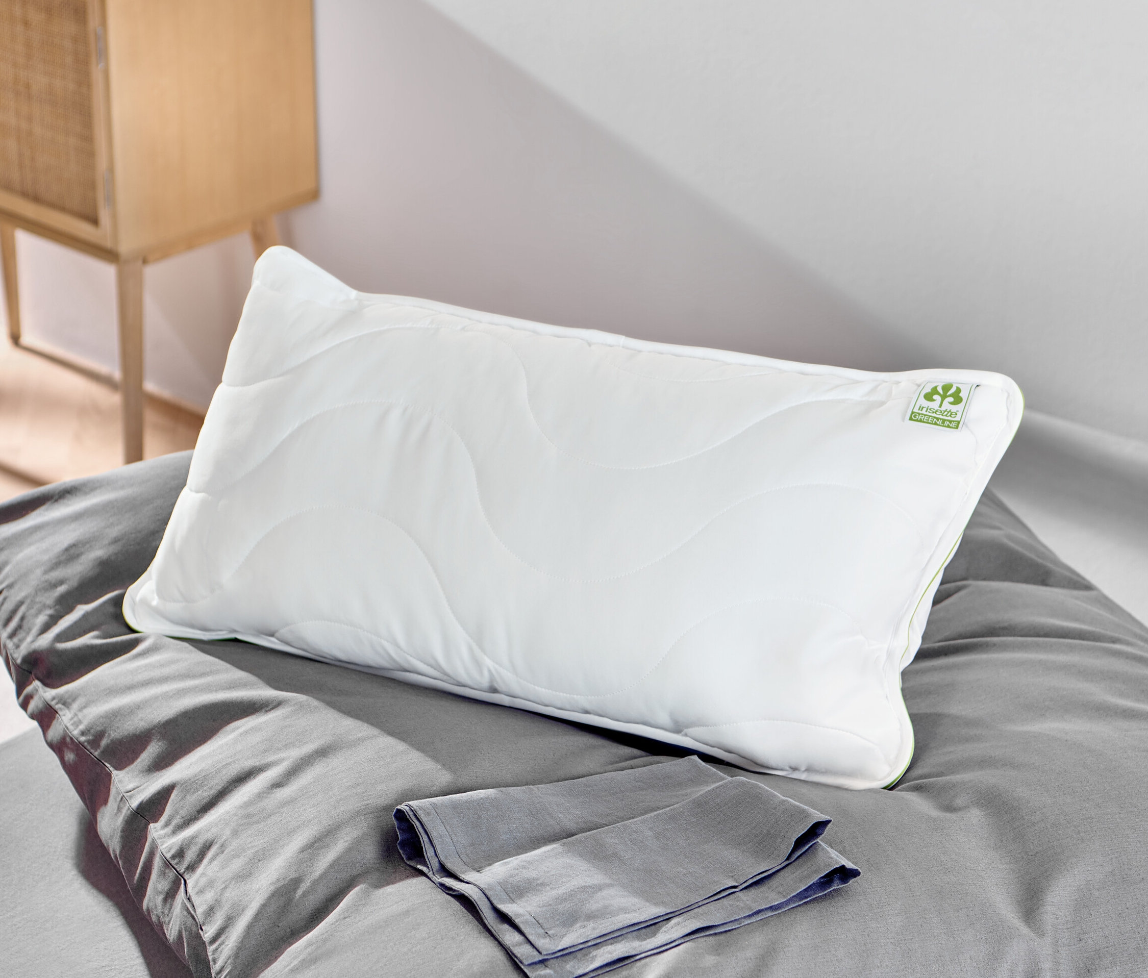 Biała poduszka irisette® greenline o wymiarach ok. 80 x 40 cm leży na szarej pościeli obok szarego ręcznika.