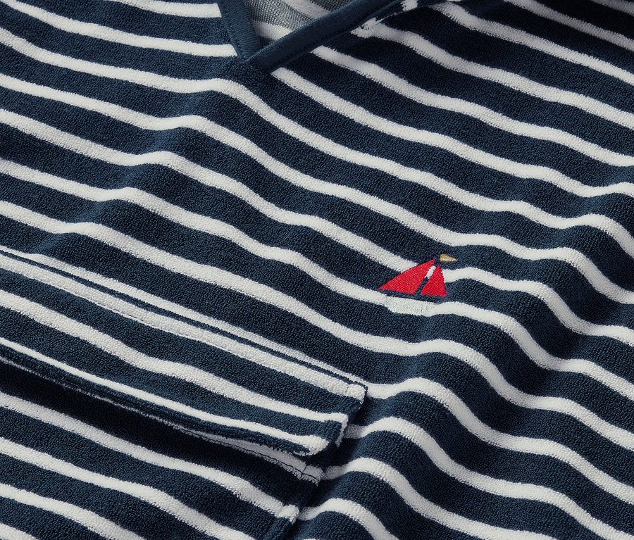 Zbliżenie na ponczo z tkaniny frotté, wybrzeże, w niebiesko-białe paski, z małym czerwonym haftem przedstawiającym żaglówkę.