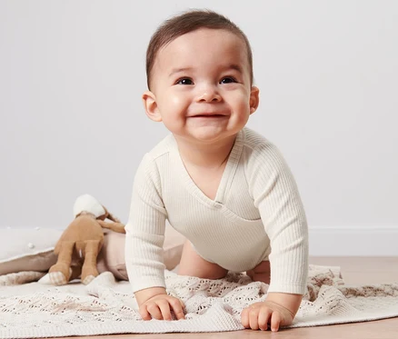 Uśmiechnięte dziecko w body z modalem raczkuje po beżowym kocu obok pluszowego króliczka.