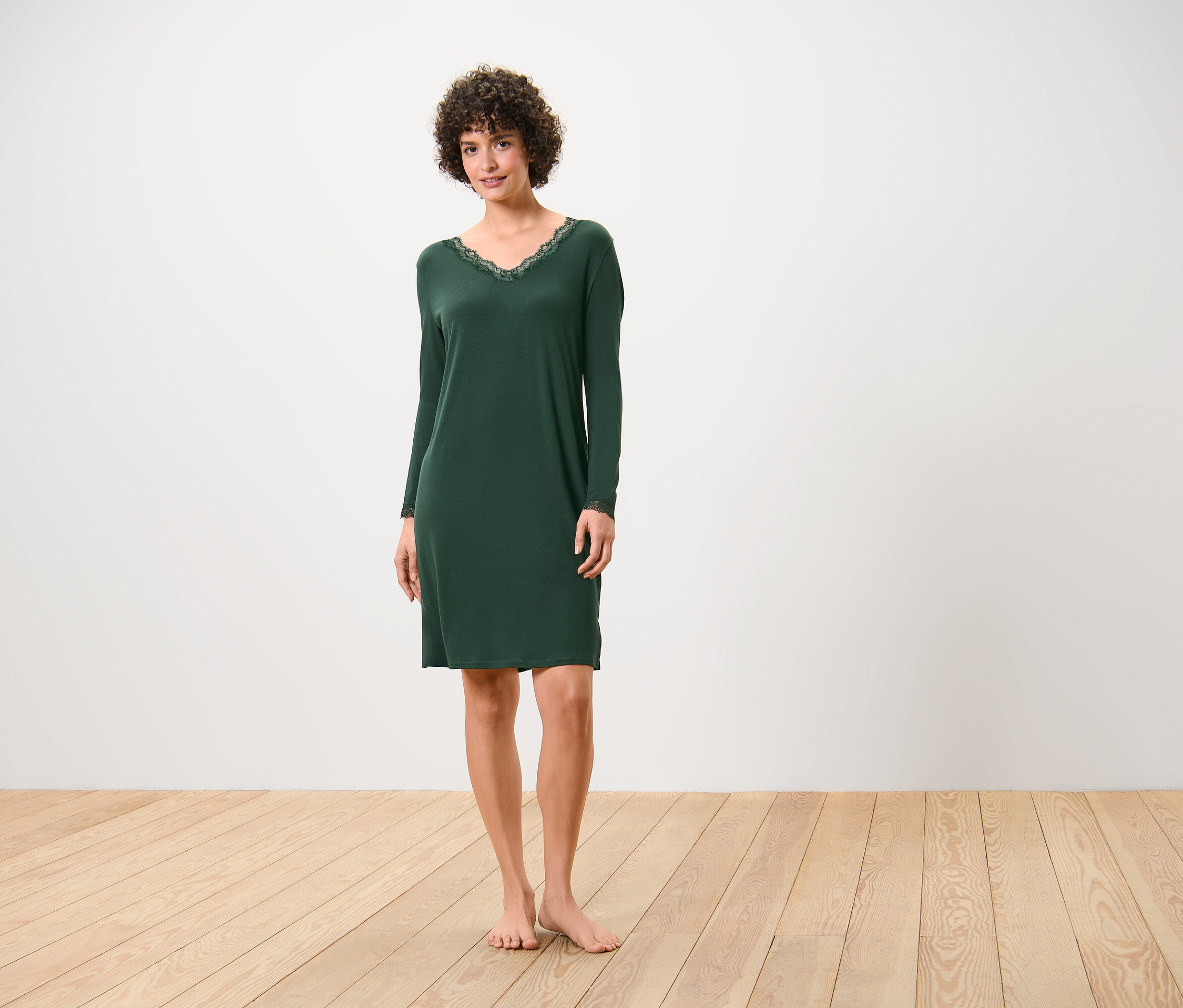 Kobieta stoi boso na drewnianej podłodze, ubrana w ciemnozieloną koszulę nocną z długimi rękawami i koronką przy dekolcie.