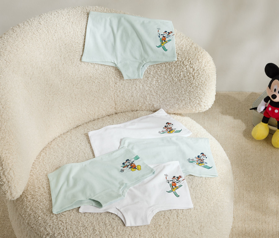 Na puszystym fotelu leżą cztery pary majtek dziecięcych z obrazkiem Myszki Miki na nartach, a obok fotela siedzi pluszowa Myszka Miki.