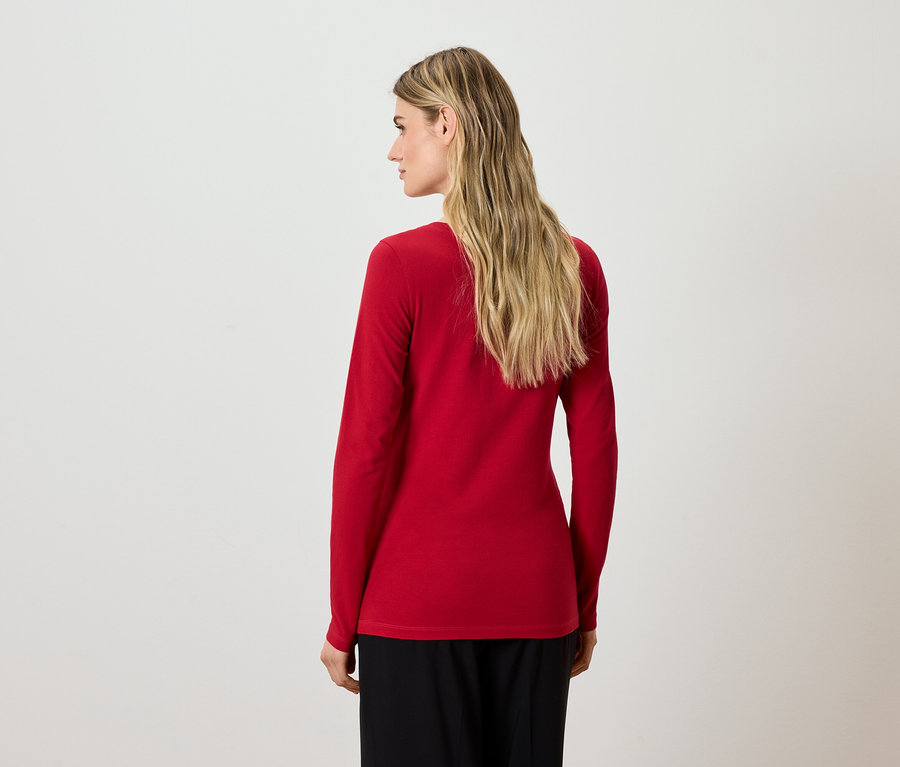 Blondynka stoi tyłem, ubrana w czerwoną bluzkę z długim rękawem i czarne spodnie.