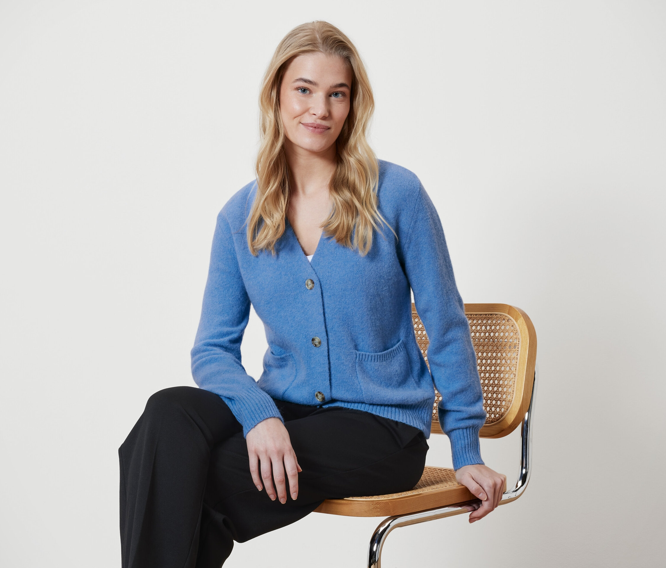 Blondynka siedzi na krześle w niebieskim swetrze i czarnych spodniach.