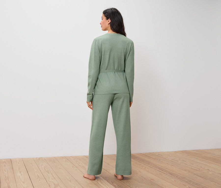 Kobieta w zielonym swetrze i spodniach stoi tyłem do widza na drewnianej podłodze.