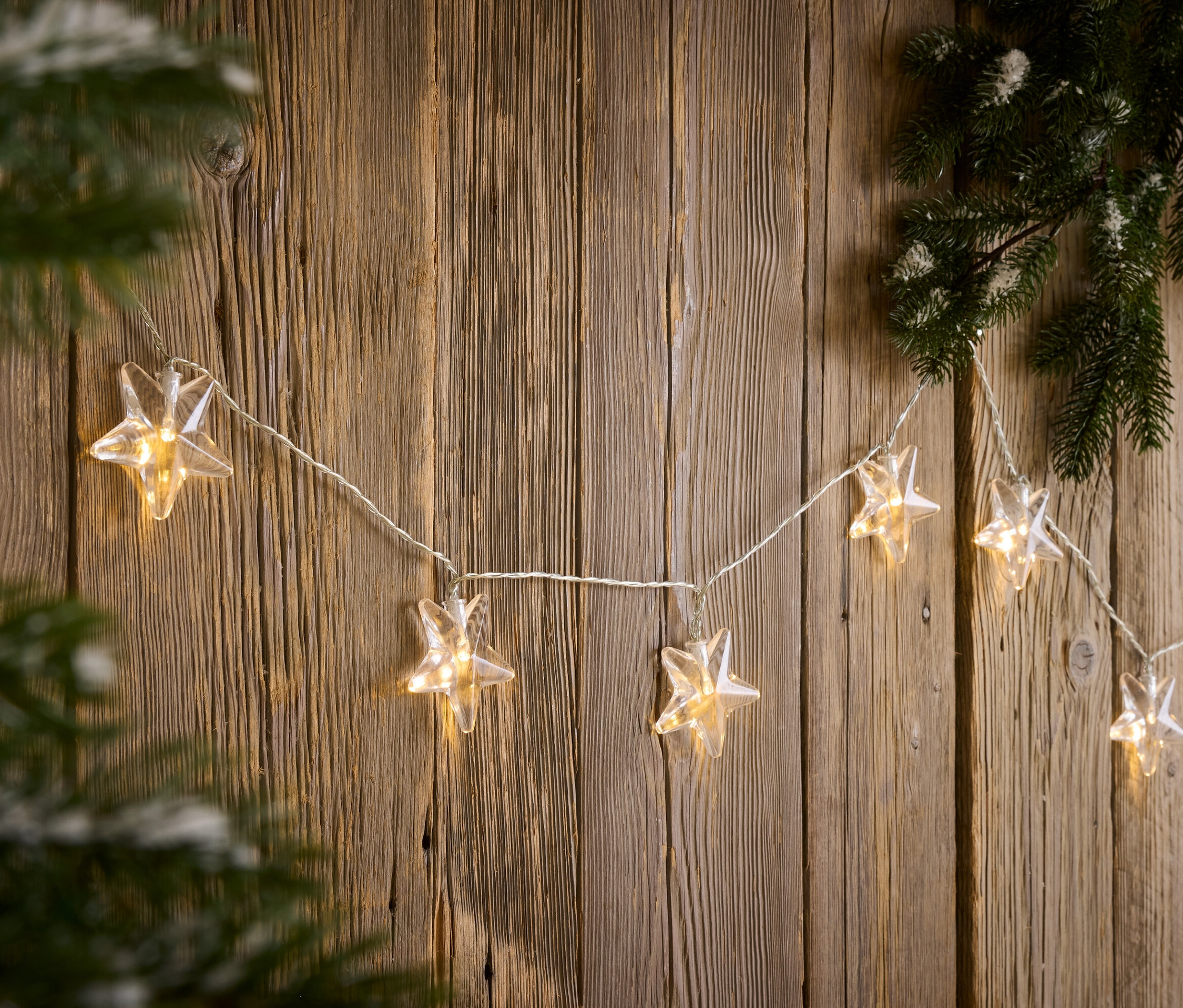 Łańcuch światełek w kształcie gwiazdy wisi na drewnianej ścianie obok gałęzi sosny.