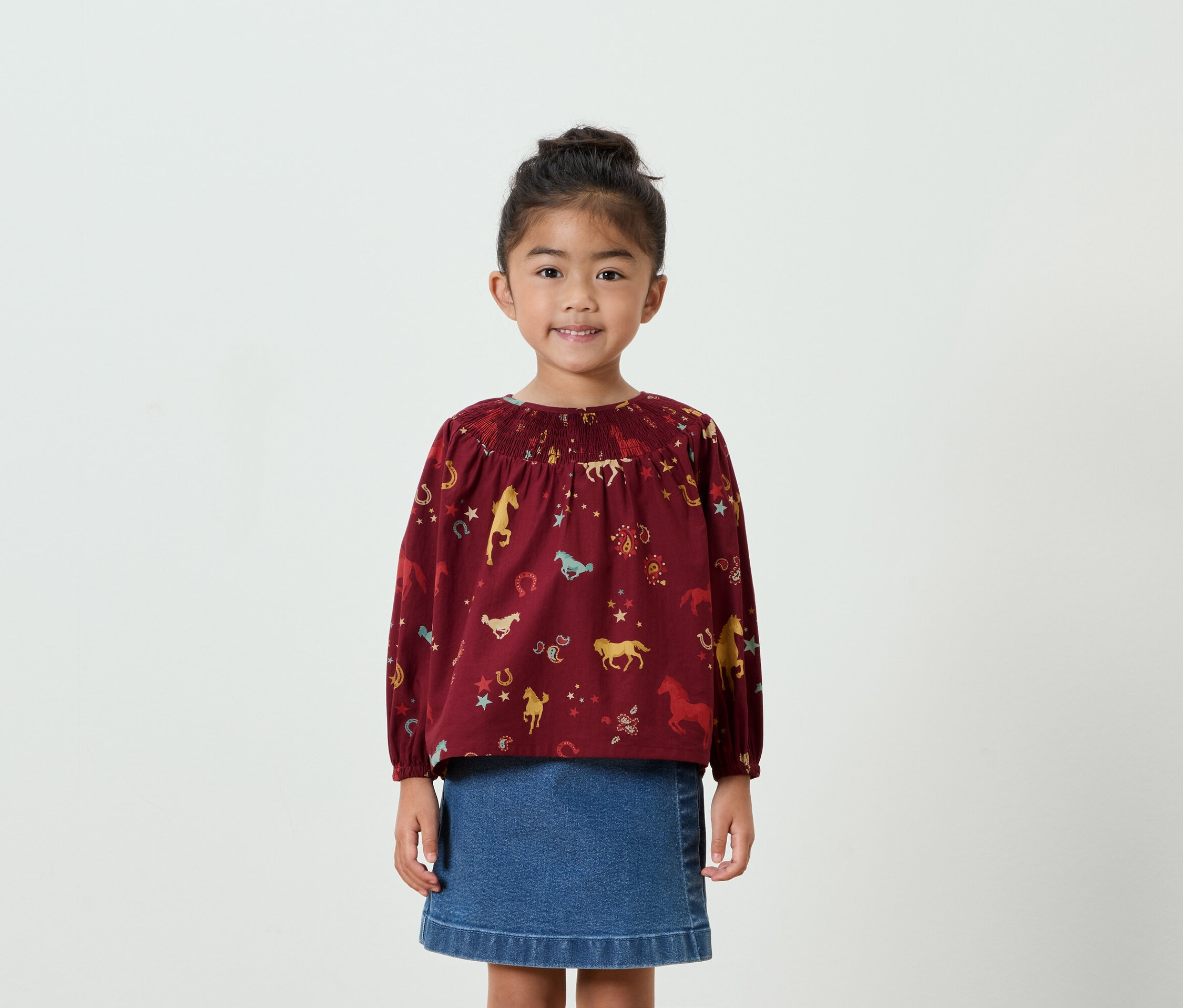 Uśmiechnięta dziewczynka pozuje w bordowej bluzce w konie i dżinsowej spódnicy.