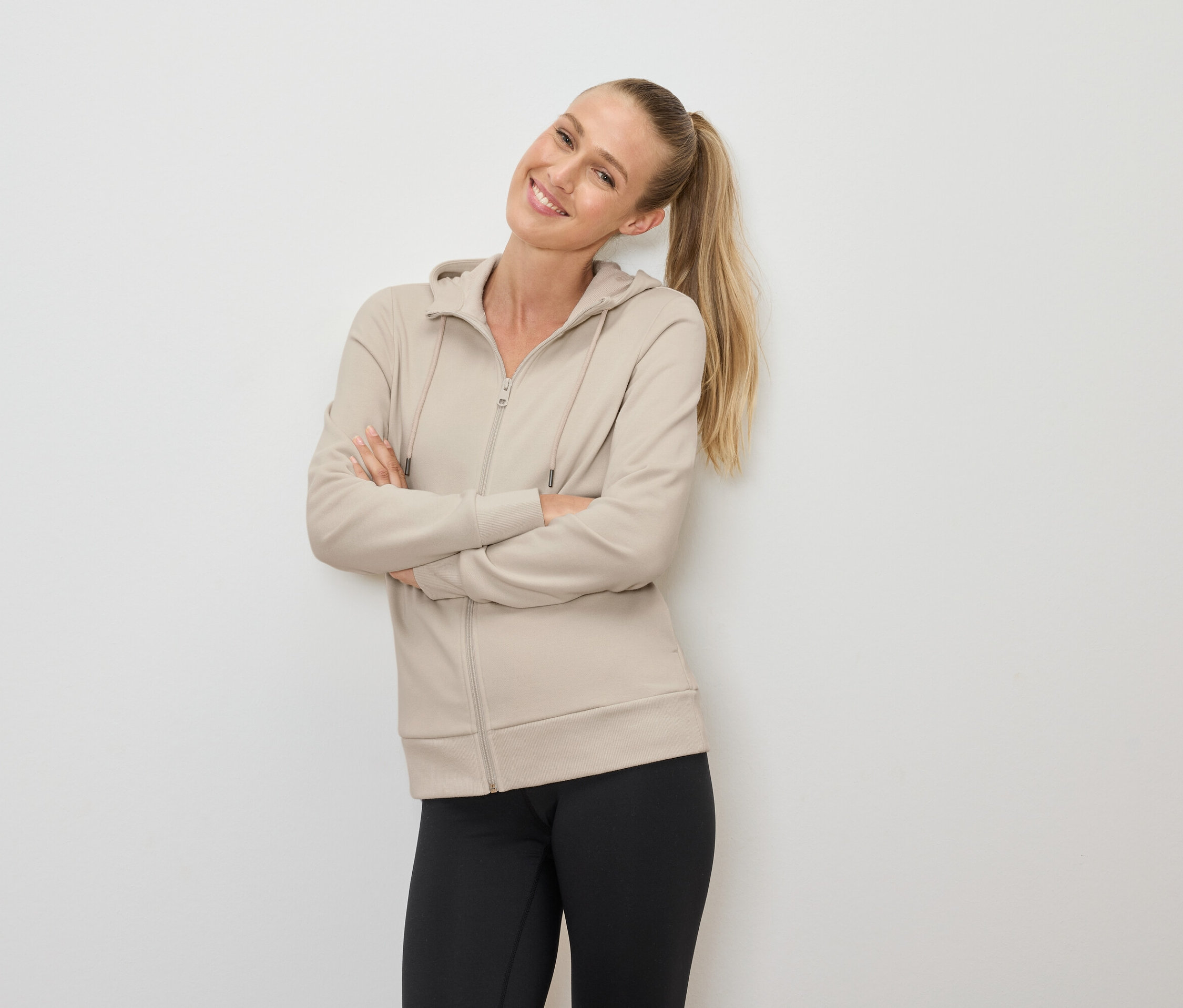 Uśmiechnięta blondynka w kucyku pozuje ze skrzyżowanymi ramionami, ubrana w beżową bluzę z kapturem na zamek i czarne legginsy.