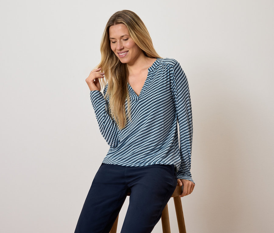 Kobieta siedzi na stołku, ubrana w niebiesko-białą bluzkę w paski z długim rękawem i ciemnoniebieskie spodnie.