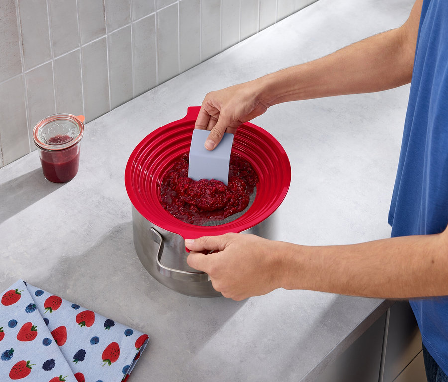 Mężczyzna przeciera dżem malinowy przez 2 w 1: sitko do przesiewania i przecierania do garnka za pomocą szarej szpatułki.