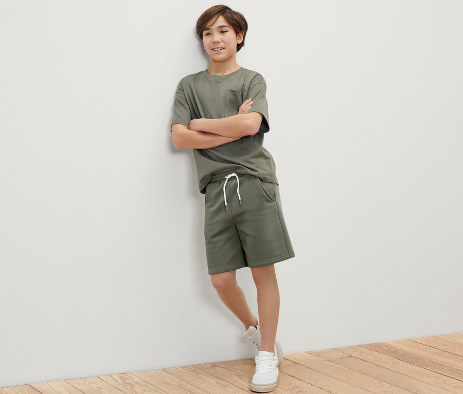 Chłopiec w khaki komplecie: T-shirt i szorty dresowe, 2 pary, stoi oparty o ścianę.