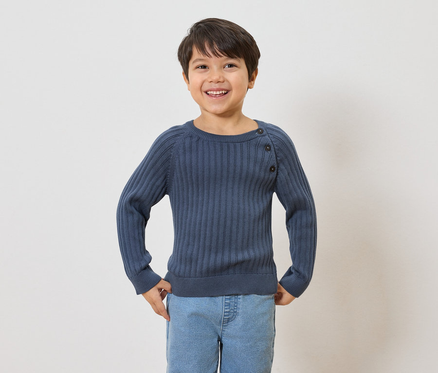 Uśmiechnięty chłopiec z brązowymi włosami ma na sobie granatowy sweter w prążki z guzikami i jasnoniebieskie dżinsy.