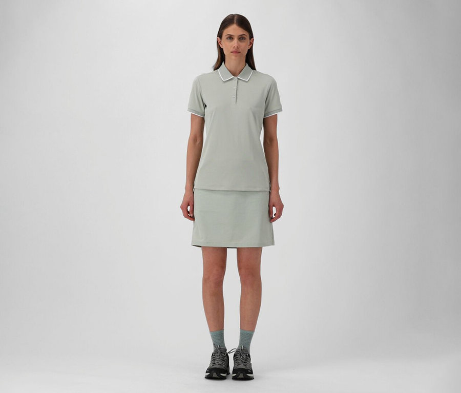 Kobieta w spódnicy outdoorowej CMP i pistacjowej koszulce polo CMP z elastycznej dzianiny piqué, szare skarpetki i buty trekkingowe.